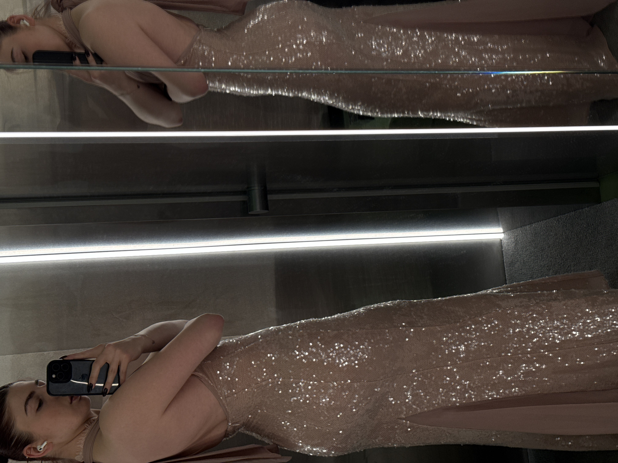 A person is reflected in a mirror, taking a selfie with a smartphone. The person is wearing a sequined dress. The environment includes artificial lighting. The location is possibly an elevator. There is no indication that the image was taken in Moscow, Russia.