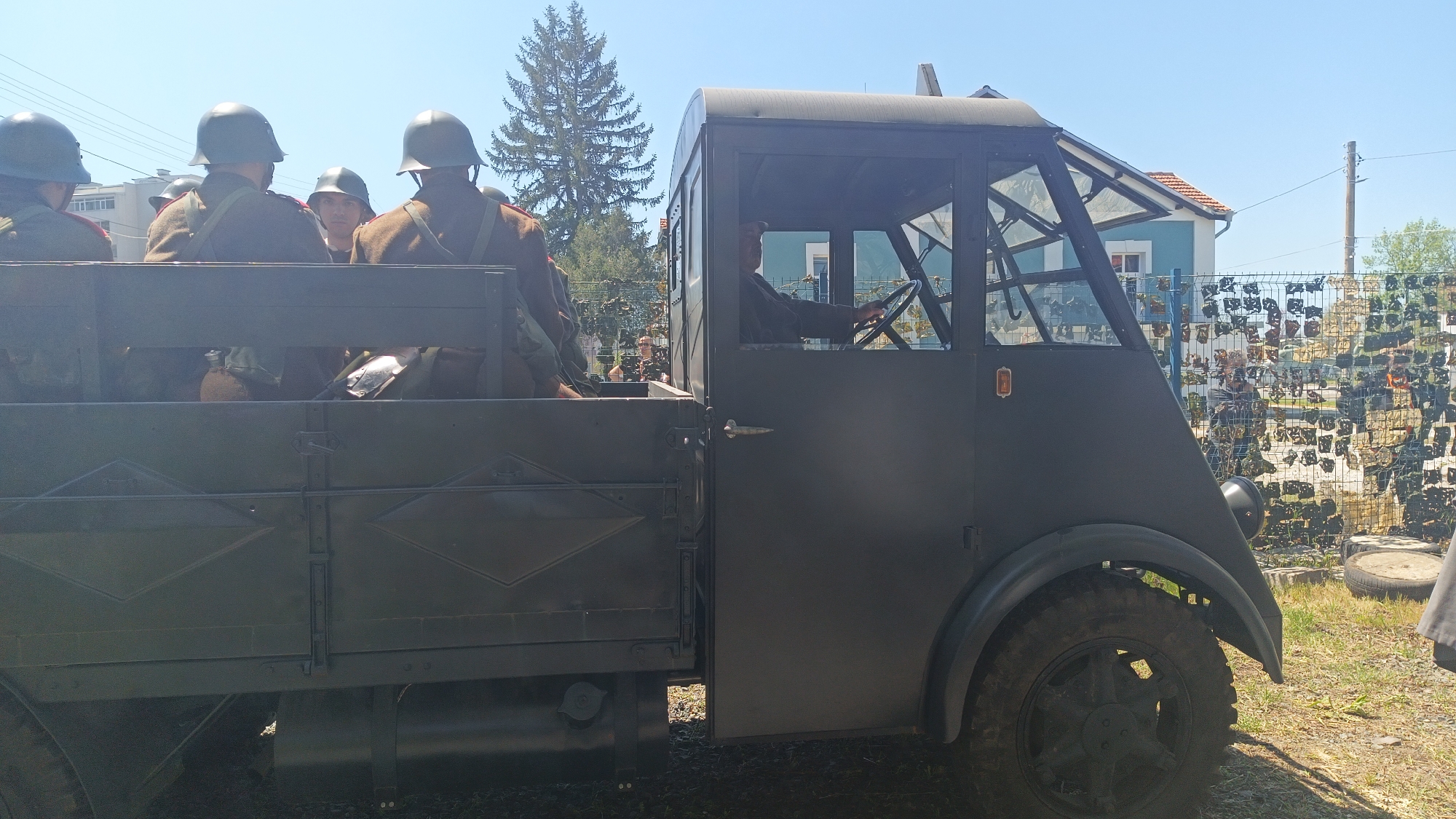 The image shows an antique, dark-colored military truck, possibly in the city of Yambol, Bulgaria. The truck bed contains soldiers wearing helmets and uniforms. A driver is visible behind the wheel. In the background, a building is partially visible, and a fence with camouflage netting is present. The sky is blue. The setting appears to be outdoors in daylight.