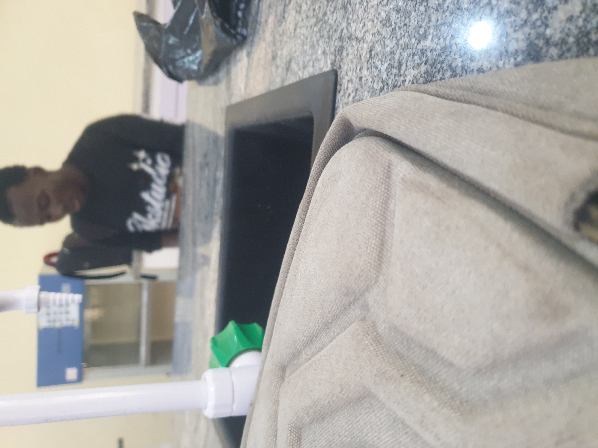 The image depicts a close-up view of a light-colored textured fabric, likely a bag, occupying the foreground. Behind the fabric, a black sink is visible on a speckled grey countertop. A white faucet with a green handle is positioned above the sink. In the mid-ground, a blurred figure of a young Black male wearing a black t-shirt with white text is seated at a counter. The background shows a blue laboratory cabinet and a yellow wall. A black plastic bag is also visible on the countertop to the right of the sink. The scene appears to be in a laboratory or a similar institutional setting.