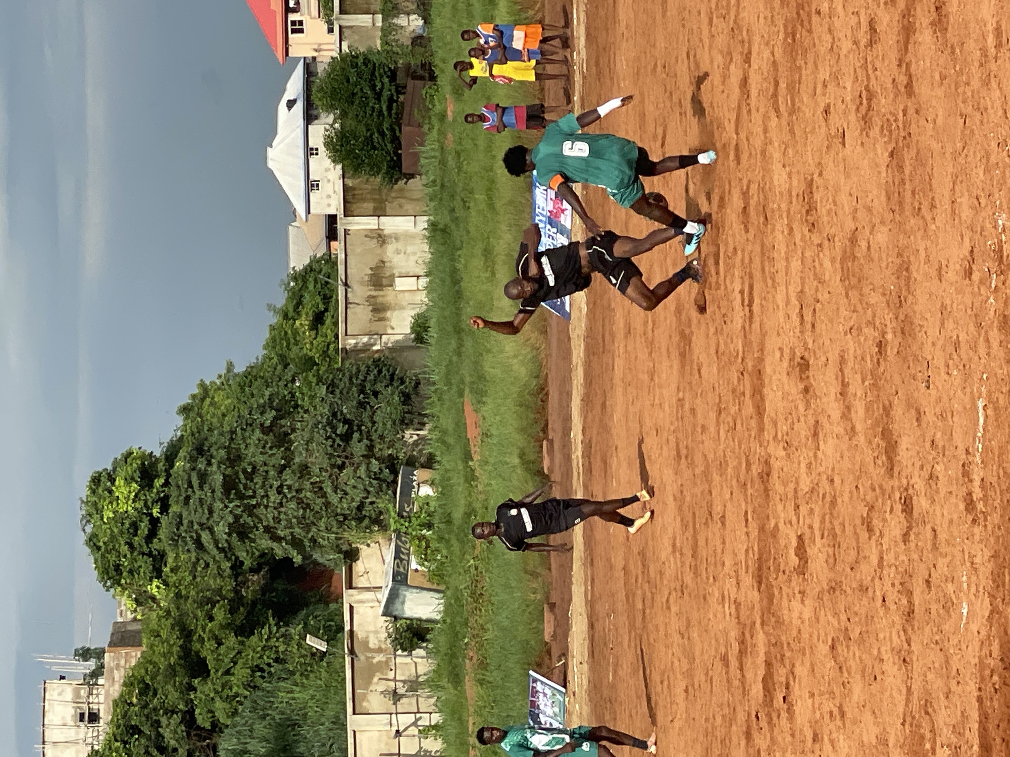 This is a medium shot, taken from a low angle, of a soccer game being played on a dirt field under a cloudy sky. In the foreground, two young men are actively engaged in a play, one in a black jersey and shorts, the other in a green jersey with the number 6 on the back. The man in black is mid-air, jumping with his arms raised as he attempts to block the ball, while the man in green is on the ground, kicking the ball.
To the left of the main action, another young man in a green jersey is visible, standing and looking towards the play. In the background, a group of young men in colorful athletic wear stand watching the game. The field is bordered by overgrown grass and a concrete wall. Behind the wall, houses with red and white roofs are visible, along with trees and a building under construction. The overall impression is of a local sporting event taking place in a developing urban environment.