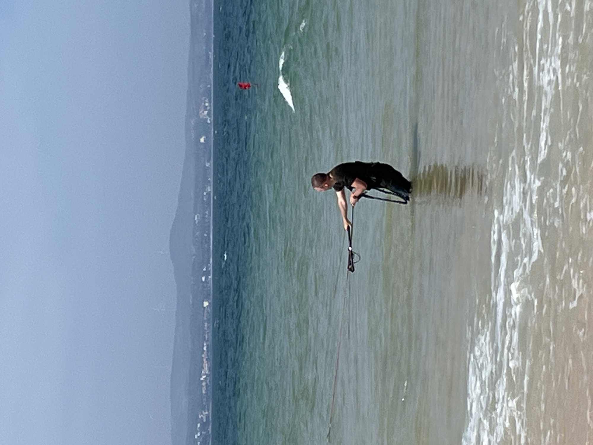 In the city of Phan Thiết, Vietnam, a man stands in the shallow, choppy turquoise waters of the sea, facing away from the viewer. He is wearing a dark t-shirt and dark pants or shorts. His hands are gripping a control bar for what appears to be a kiteboarding setup, which is extended in front of him. The water reaches his mid-thighs. In the background, the sea stretches to the horizon, with a faint, hazy coastline dotted with buildings and distant wind turbines visible under a pale sky. A small red flag stands upright in the water to the right of the man, and a wave crests in the middle distance. The foreground shows the sandy bottom of the sea transitioning to a gentle wave break at the shoreline.