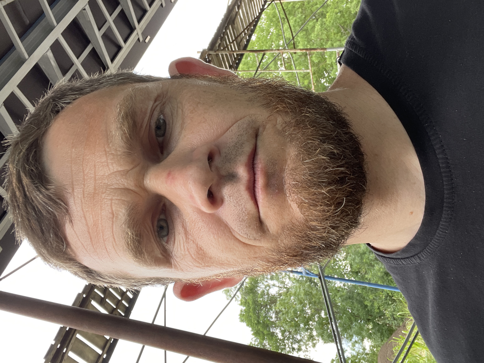 A man with light brown hair and a beard is taking a selfie outdoors. He is wearing a black t-shirt. The background shows some construction elements and trees, suggesting an outdoor or semi-outdoor setting. The lighting indicates it is daytime. The man's expression is neutral. There is no indication of the specific location within Krasnodar, Russia.
