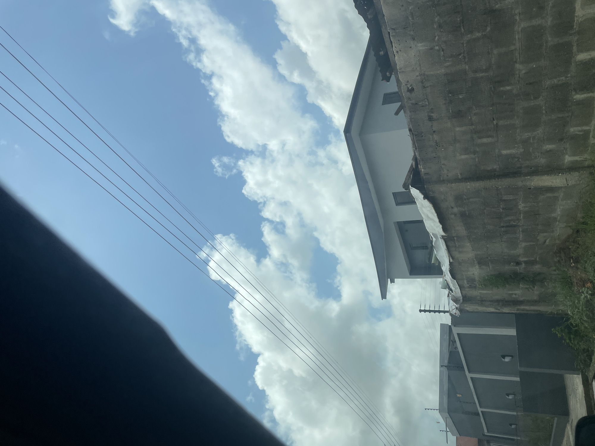 The image shows a view of the sky with scattered clouds and a few overhead power lines. Buildings are visible on the right side of the frame, partially obscured by a concrete block wall. A portion of what appears to be a car's interior or a dark surface is visible at the bottom left corner. Based on the prompt, the location is Ado, Nigeria, suggesting a neighborhood or residential area, but without further details, the specifics of the location within the city cannot be determined. No people or interactions are visible.