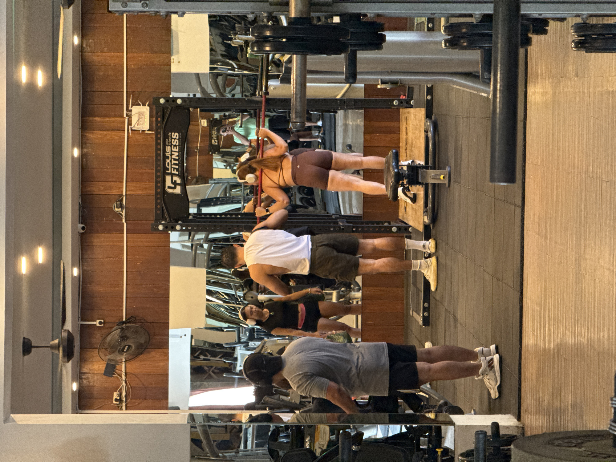 The image shows the interior of Louis and Friends Fitness gym in North Kuta, Indonesia. Three people are exercising in front of a mirrored wall.
A person in a gray shirt and black shorts is doing push-ups. A second person in a white shirt and olive shorts is doing an upright row, using a machine. A third person in brown exercise clothing is doing some kind of leg work using another machine. Other people are visible in the mirror.
Behind the people are weight machines and a wooden wall with a ceiling fan. A black fitness logo is visible on one of the weight racks. The flooring appears to be a mix of materials. The lighting is bright, with a combination of natural and artificial sources.