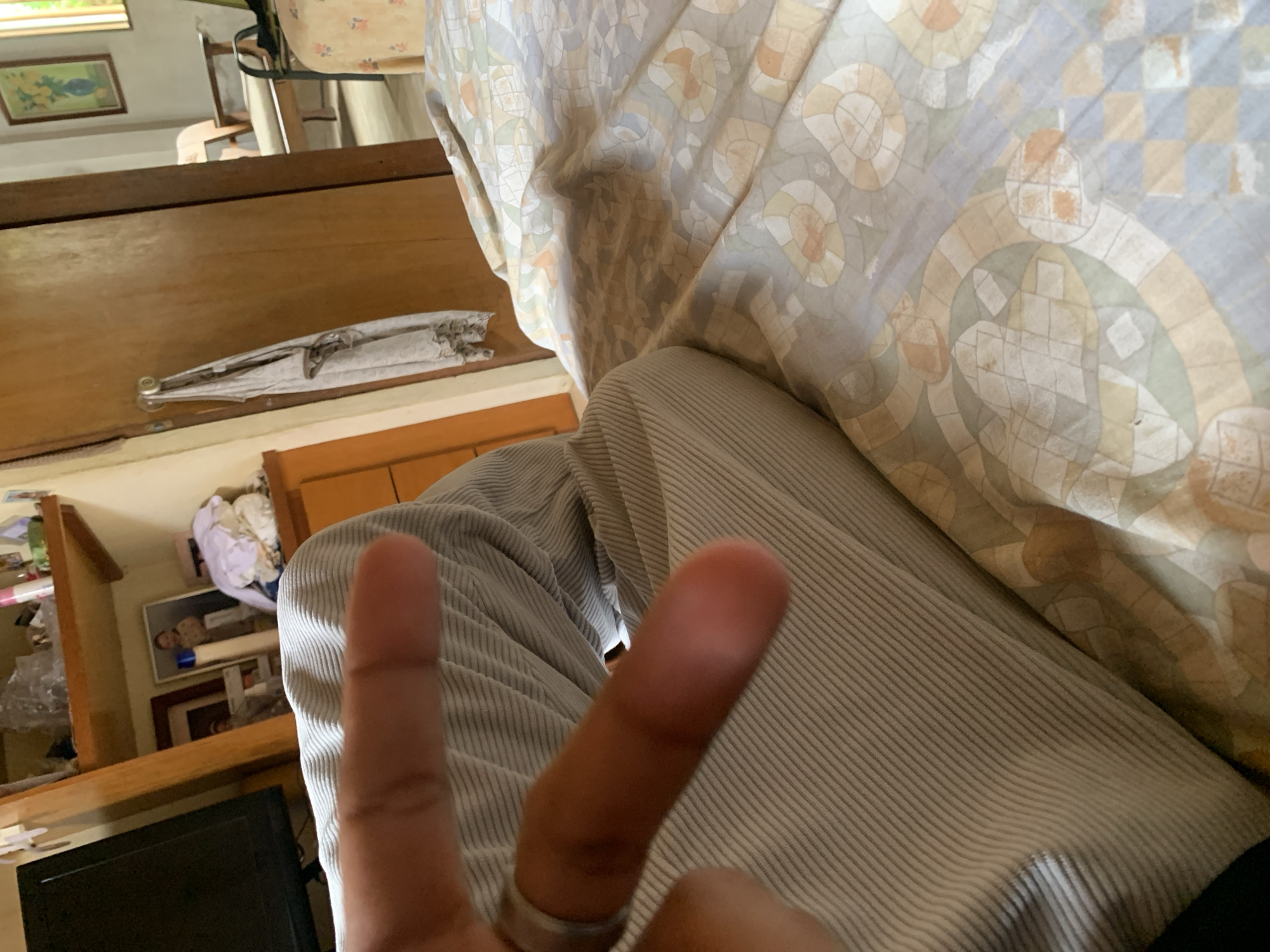 A person's hand with two fingers extended in a peace sign gesture is positioned in the foreground of the image, obscuring a portion of their lap which is covered in light-colored, pinstriped fabric. The background reveals a bedroom setting. To the left, a television is visible on a shelf unit. The shelves above contain various items, including what appears to be a stack of laundry and framed pictures. Further back, a wooden door is slightly ajar, with a piece of fabric hanging from a hook on it. Beyond the door, glimpses of another room show furniture, including a chair and a bed with a patterned comforter. The overall impression is of an interior domestic space in what is likely a residential area of Santa Ana, Venezuela.