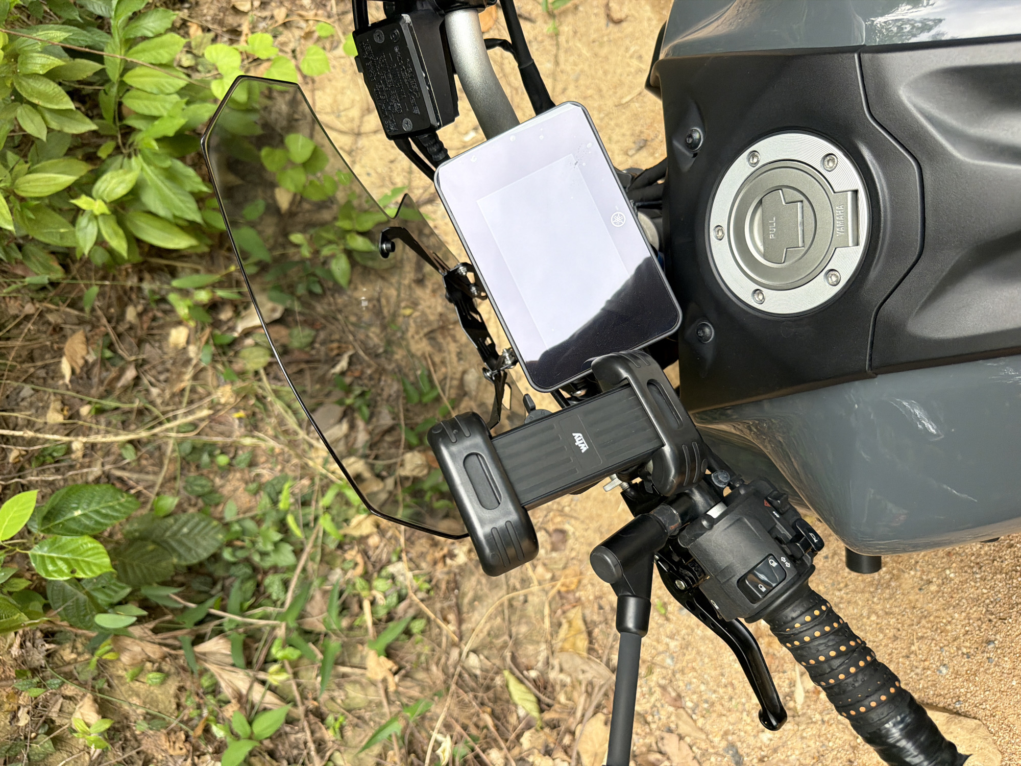 The image shows a close-up, overhead view of the handlebars and dashboard of a grey Yamaha motorcycle. The motorcycle is parked outdoors on a sandy surface, with a background of lush green foliage and dry leaves. Mounted on the handlebars is a black phone holder, and to its right is the motorcycle's digital display, which is illuminated and shows a blank white screen. The fuel cap, featuring the Yamaha logo and the word "PULL," is visible on the motorcycle's tank. The scene is consistent with a parked motorcycle in a natural, outdoor setting, likely in Ko Samui, Thailand, given the presence of tropical vegetation. There are no people or animals in the frame, and no discernible activity is taking place.