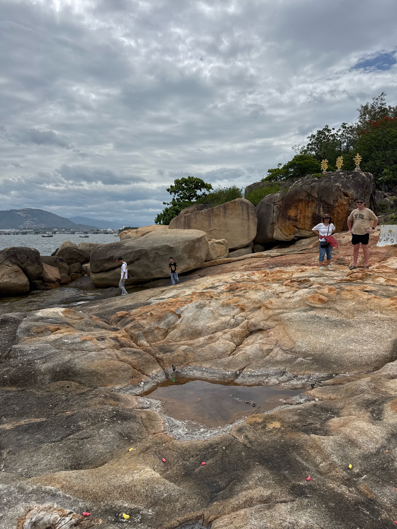This is a full shot, taken from a low angle, of a rocky shoreline with the ocean and distant mountains in the background. The sky is overcast with thick clouds. On the rocks in the foreground, there are four people. Two men and two women. A couple, in their 50s, stand in the middle of the frame, looking at the camera. The woman is wearing a white t-shirt, blue jeans, and sunglasses. The man is wearing a tan t-shirt, black shorts, and sunglasses. Two young men, one in a white t-shirt and gray pants, and the other in a black t-shirt and jeans, are walking away from the camera towards the water. The rocks are a mixture of brown and gray, with some reddish-brown hues. There is a small tide pool in the foreground. In the background, there are large boulders and some vegetation. On the largest boulder, there are three golden statues of Buddhist characters. The ocean is calm, with a few boats visible in the distance. A bridge and city skyline can be seen on the left side of the frame. The image is taken at Hon Chong Promontory in Nha Trang, Vietnam.