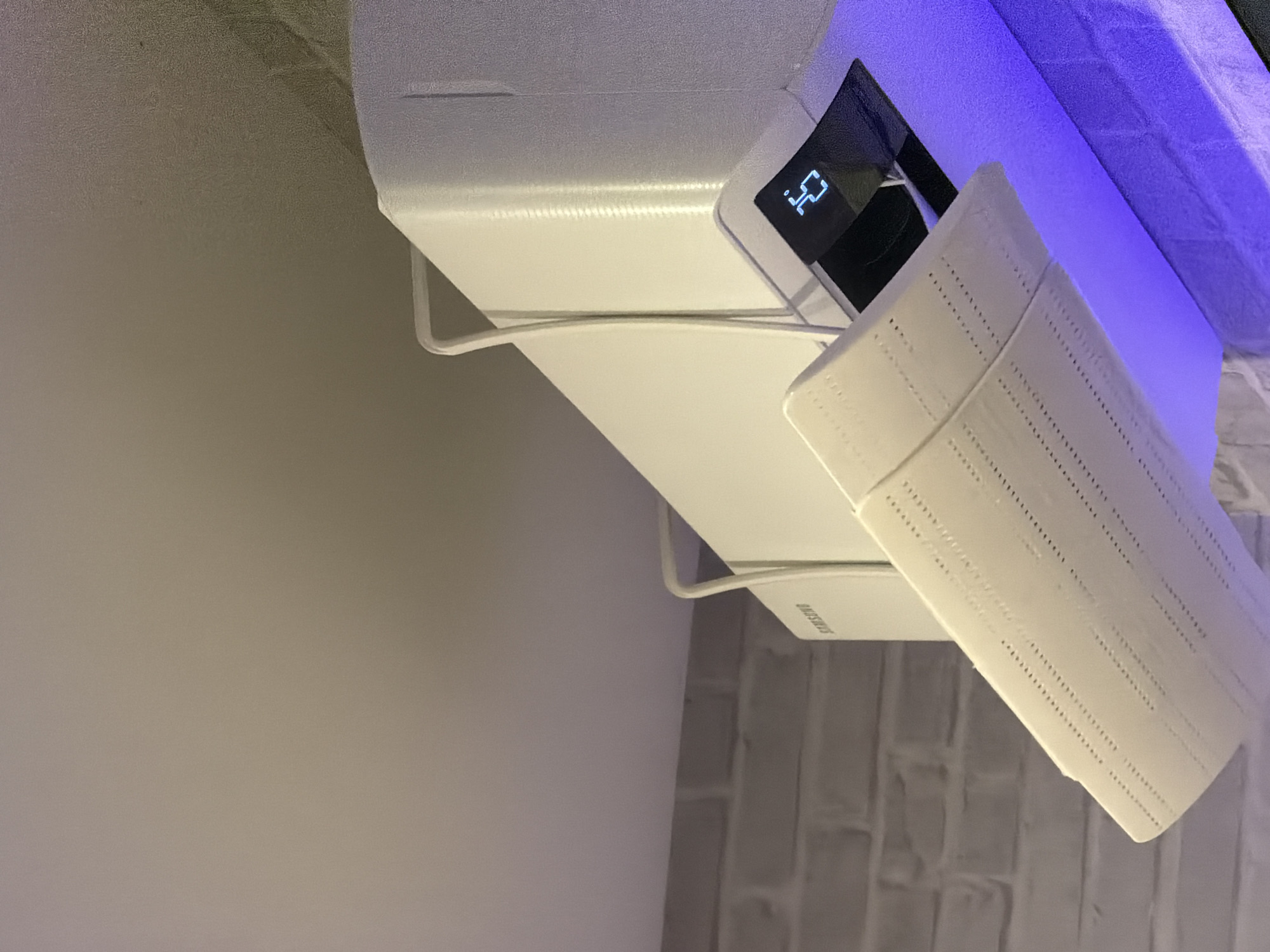 An indoor Samsung air conditioning unit is mounted on a white brick wall, partially illuminated by blue LED lighting. The unit has its vent open, and a digital display shows "25". The ceiling appears to be a textured white surface. The image is taken from a low angle looking upwards.