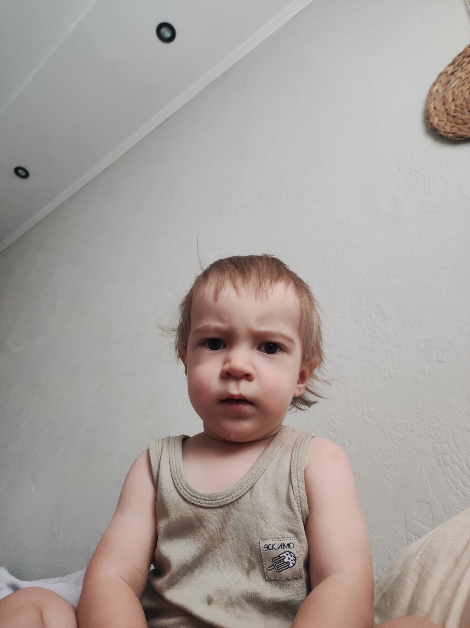 A close-up shot of a young child is positioned in the lower half of the frame, looking directly at the viewer with a furrowed brow. The child has fair skin, light brown hair with some strands standing up, and is wearing a light beige tank top with a small square patch featuring an image and Cyrillic text on the lower left.
The background is a neutral-colored wall, slightly textured. Near the top left corner, there are two recessed ceiling lights. A decorative woven item is visible in the upper right corner of the frame.
The location cannot be verified based on the provided image. The presence of Cyrillic text on the shirt suggests a possible Russian-speaking origin, but without additional contextual clues, a specific location like Chaikovsky, Russia, cannot be confirmed.