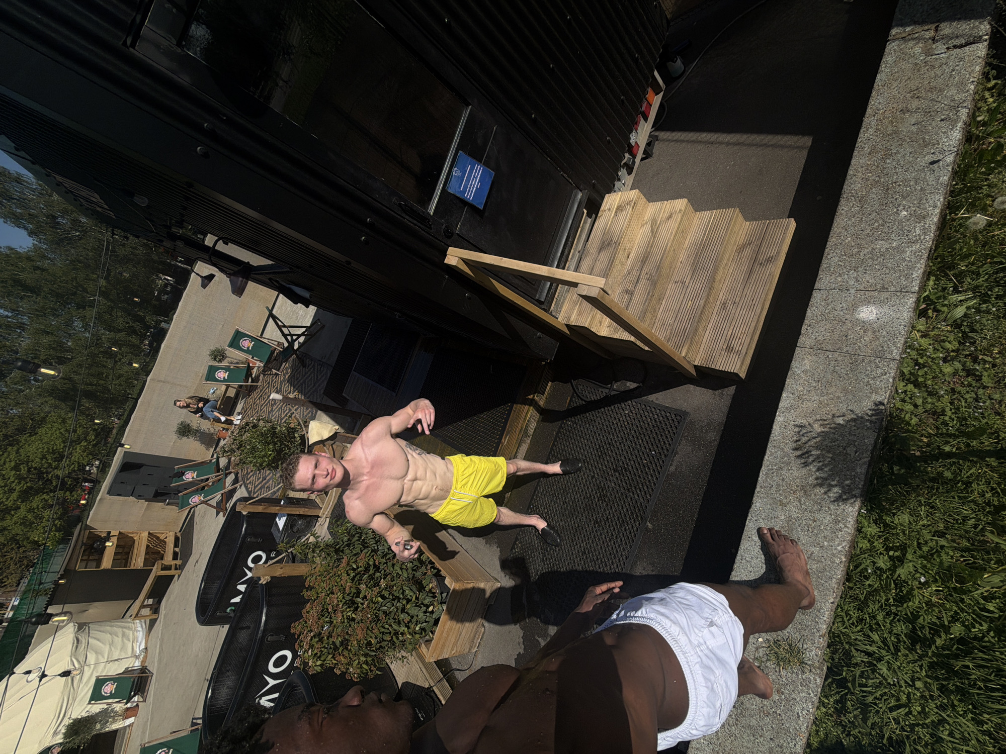 The image, taken from an elevated perspective, captures an outdoor event or wellness area under bright, direct sunlight. In the central foreground, a man with a muscular build, shirtless and wearing yellow swim shorts and dark slip-on shoes, lies on his back on a black mat. He appears wet, suggesting he may have recently used a plunge tub. He looks directly towards the camera. In the lower foreground, partially visible, is another person (likely a man) wearing white swim shorts, with dark skin.
The scene features a large, dark, corrugated metal structure, possibly a temporary building or converted shipping container, with wooden stairs leading to an entrance. Adjacent to these stairs and the mat, two black plunge tubs are visible, each displaying "MYO" in white text. A patio area in the background is furnished with several green deck chairs that feature an unidentifiable crest logo. Potted plants are strategically placed around the area, and string lights are strung overhead. In the distance, a woman is seated on one of the chairs. The ground consists of paved asphalt and concrete, bordered by a concrete slab and a grassy area. A small, illegible blue sign is affixed to the dark structure. The strong sunlight creates distinct shadows, indicating a clear, sunny day in what is specified as London, UK. The overall atmosphere suggests a relaxed, recreational, or recovery setting at an outdoor venue.