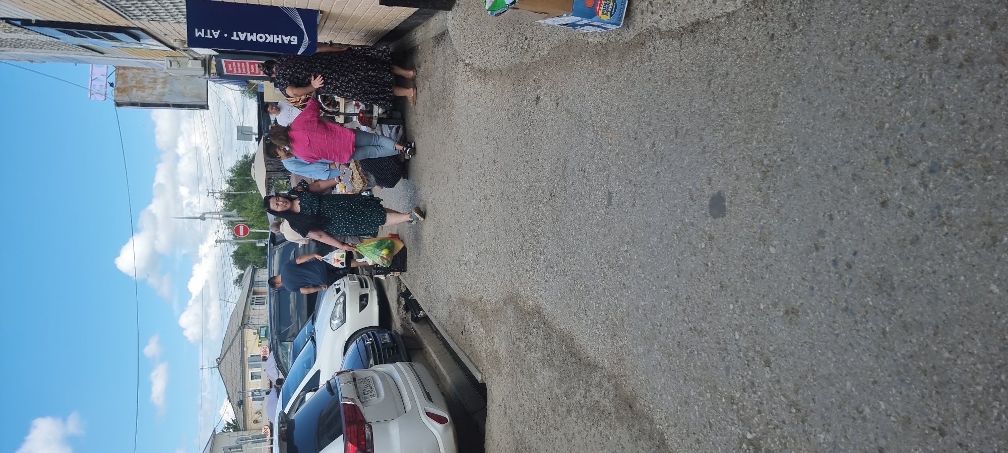A street-level view captures a group of people walking along a paved sidewalk next to parked cars and buildings. The sky above is blue with scattered white clouds. The people, appearing to be of Asian ethnicity, are dressed in casual clothing. Some are carrying shopping bags. One woman in a dark, polka-dot dress is walking towards the camera, holding a bag with some fruit and a carton of eggs. Another woman in a bright pink shirt is seen further back. In the background, a white SUV is parked, followed by other vehicles. The buildings appear to be commercial, with a sign for "BANKOMAT ATM" visible on one of them. The overall scene suggests a busy marketplace or commercial area in Aktobe, Kazakhstan.