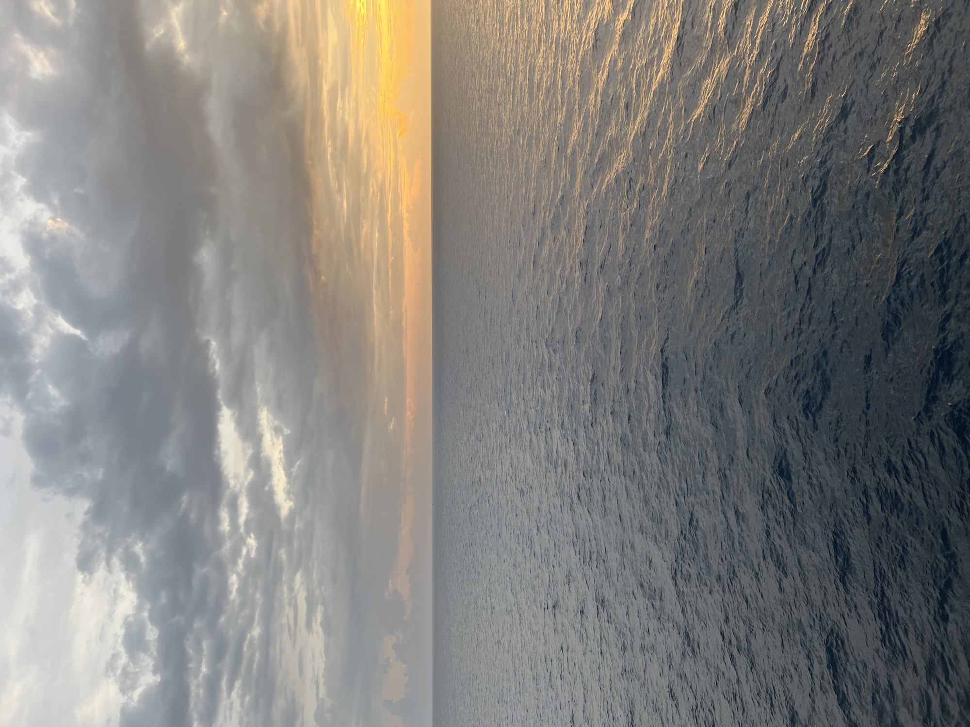The image depicts a wide expanse of the ocean under a cloudy sky, with the sun setting or rising on the horizon, casting a warm glow. The water is a deep blue-gray, with small waves creating ripples on its surface. The sky is a mix of dark gray clouds towards the top and lighter, yellowish hues near the horizon, suggesting either sunrise or sunset. There are no discernable landmarks or structures visible, placing the scene in an open marine environment, far from any urban setting. The location is therefore unknown, and no specific details about people, subjects, objects, actions, or interactions can be provided.