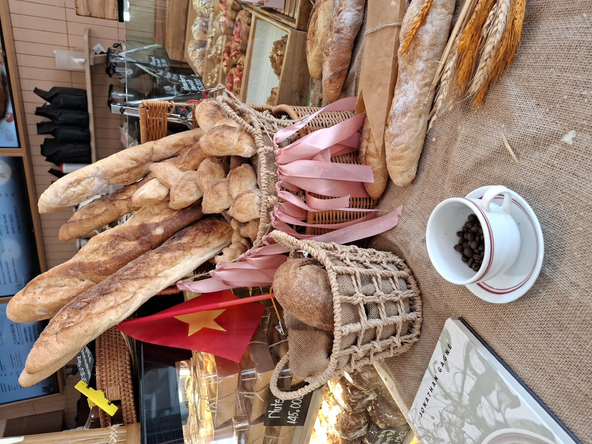 A close-up, eye-level shot depicts a collection of baked goods, primarily baguettes and small round loaves, arranged on a burlap cloth in what appears to be a bakery. The baguettes, some dusted with flour, are the most prominent items, leaning against each other in a wicker basket adorned with pink ribbons. A small Vietnamese flag, red with a yellow star, is positioned in front of the baguettes. To the left, a small woven basket holds two round loaves, one partially visible and wrapped in burlap. A price tag for "Nuts 145.000" is visible in the background. To the lower left, a book titled "JONATHAN GAGNE" rests on the burlap. A white teacup and saucer, with red trim, sit to the right of the book, filled with dark coffee beans. Several stalks of dried wheat are arranged to the right of the teacup. In the background, behind the baked goods, are display cases containing more pastries and breads, suggesting a commercial setting. The overall impression is one of a curated display of fresh bread and coffee, likely within a cafe or bakery in Nha Trang, Vietnam, given the presence of the Vietnamese flag. There are no people visible in the frame.