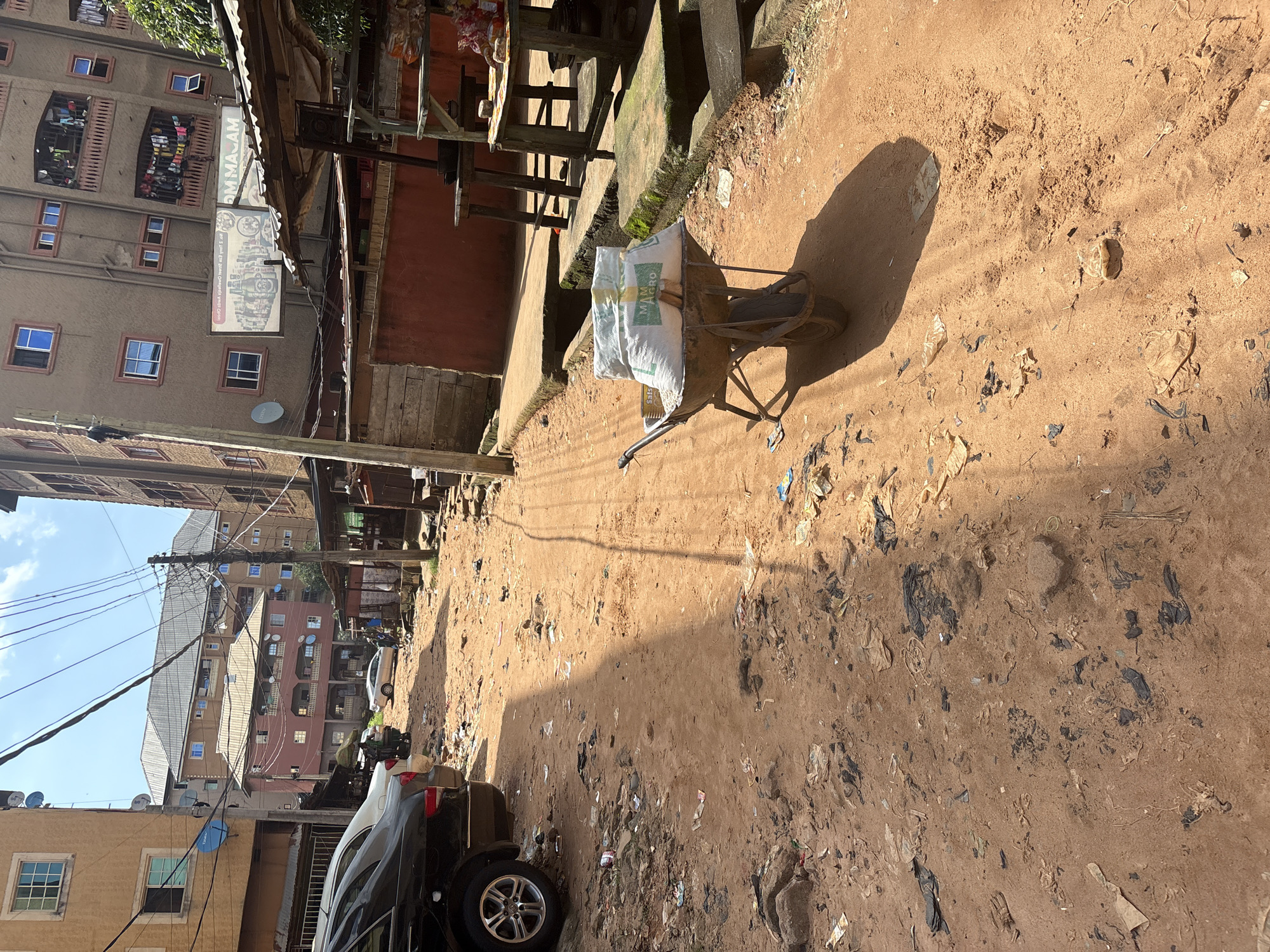 The image depicts a sun-drenched, dusty street in Nkpor, Nigeria, characterized by its unpaved, reddish-brown dirt road strewn with litter, including plastic bags and paper. The environment is densely populated with multi-story buildings, some appearing residential and others commercial, with satellite dishes visible on their rooftops. Overhead, a complex network of electrical wires crisscrosses the sky. In the foreground, a wheelbarrow loaded with sacks, one of which is labeled "MAM AGRO," stands prominently, suggesting ongoing or recent activity related to the transport of goods. Parked on the left side of the road is a dark-colored sedan. Further down the street, a few individuals can be observed, some near a small market stall. The overall scene conveys a sense of urban life in a developing area, with elements of both commerce and the challenges of maintaining cleanliness.