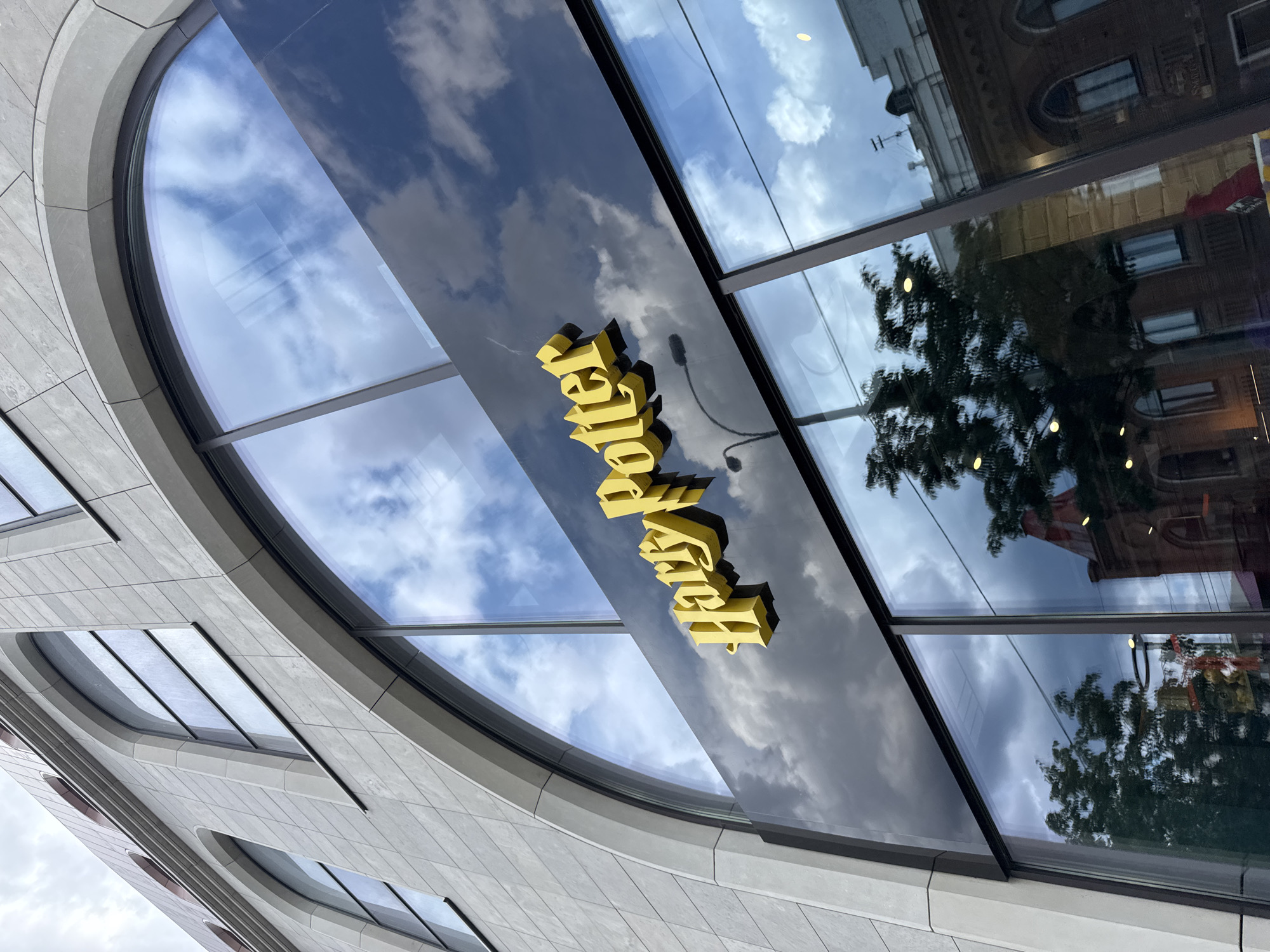 The image shows a close-up, angled view of a building's facade, featuring a large, curved, dark reflective glass window. The glass reflects a sky filled with dramatic, puffy white clouds against a blue sky. In the reflection, older buildings with arched windows and architectural details are visible, as well as the silhouette of trees.
Affixed to the reflective glass are raised, three-dimensional yellow letters that spell out "Harry Potter" in a stylized font. The letters are illuminated and catch the light, contrasting with the dark glass. The building itself appears modern, with textured stone panels and curved window elements.
The time of day is difficult to determine precisely due to the reflections and angle, but the bright sky suggests daytime. The weather appears to be clear with significant cloud cover. The presence of the "Harry Potter" branding strongly suggests this is a store or attraction related to the popular franchise. The location context of Yekaterinburg, Russia, implies this is a specific establishment within that city. There are no people visible in the frame.