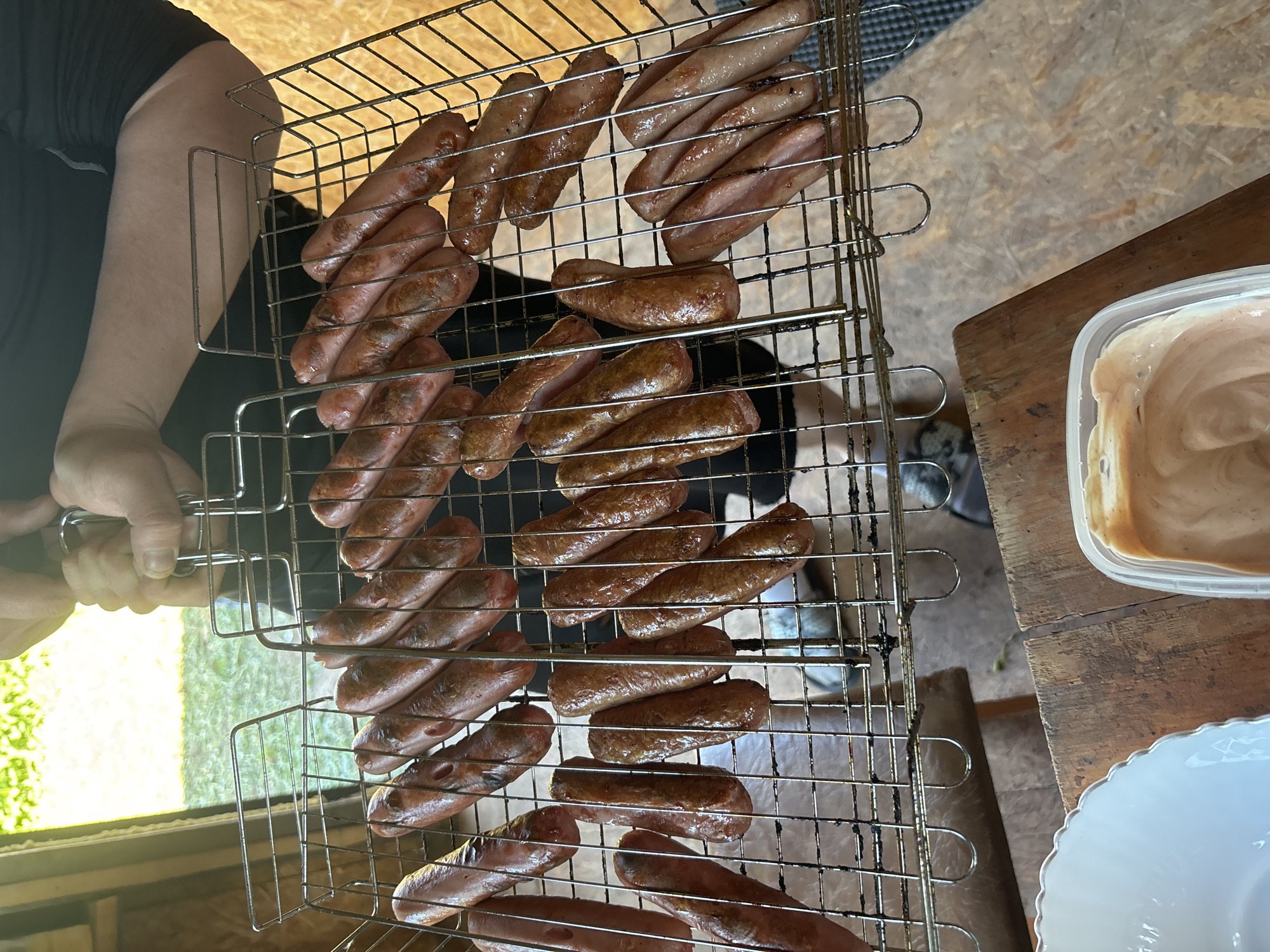 A person is holding a grill with many sausages. The sausages are evenly spaced and cooked. There is a plastic container with light brown dip and a white plate in the frame. The location is not discernable.