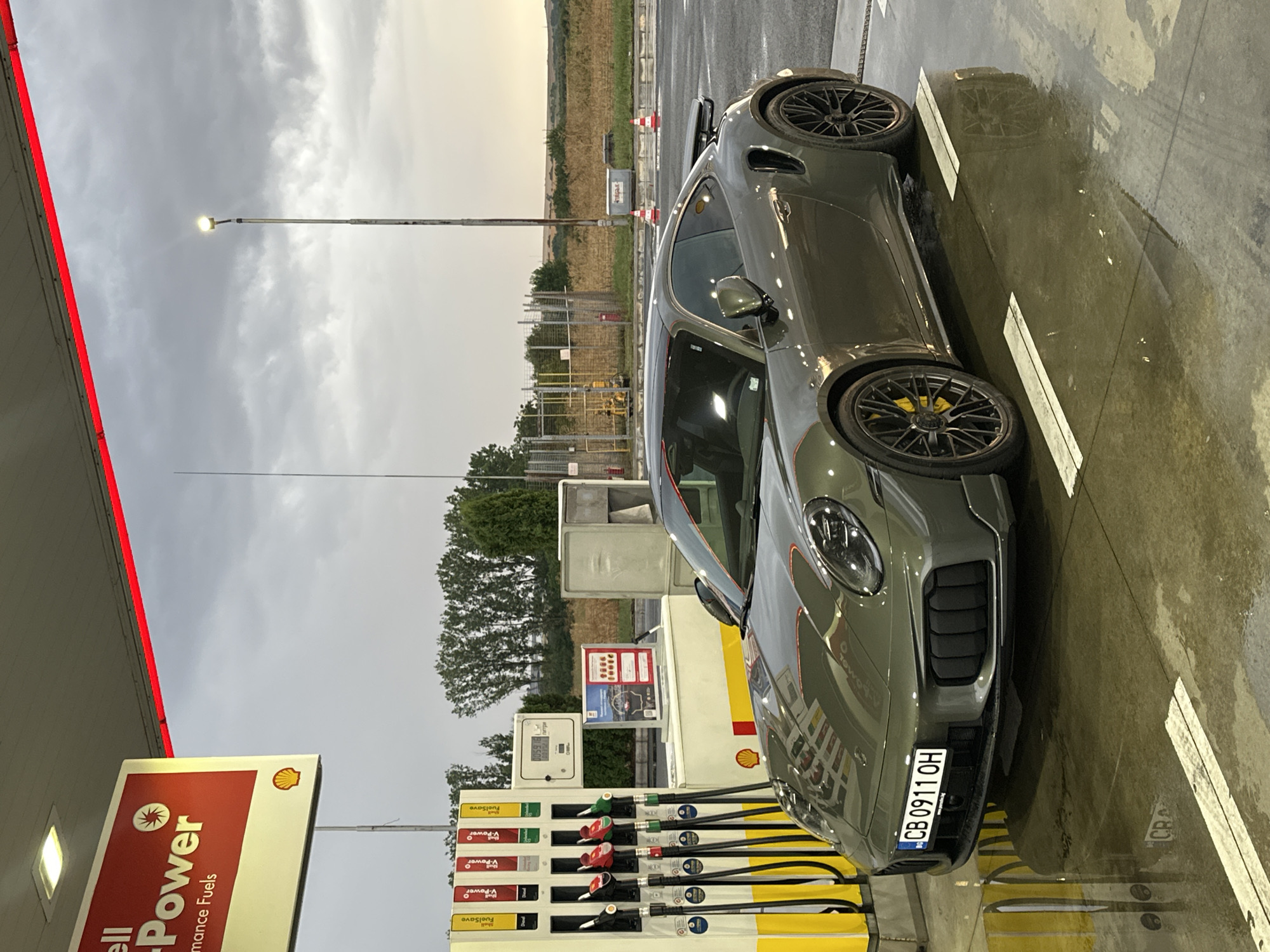 A grey Porsche 911 Turbo S is parked at a Shell gas station under a cloudy sky. The car, with its distinctive spoiler and yellow brake calipers, is positioned in front of a fuel pump island. The license plate reads "CB 0911 OH". The ground is wet, reflecting the car and the surroundings. In the background, there are trees, a street light, and a field. The Shell sign indicates "V-Power Performance Fuels." The image appears to be taken in Drazhevo, Bulgaria, given the license plate format and the surrounding landscape.