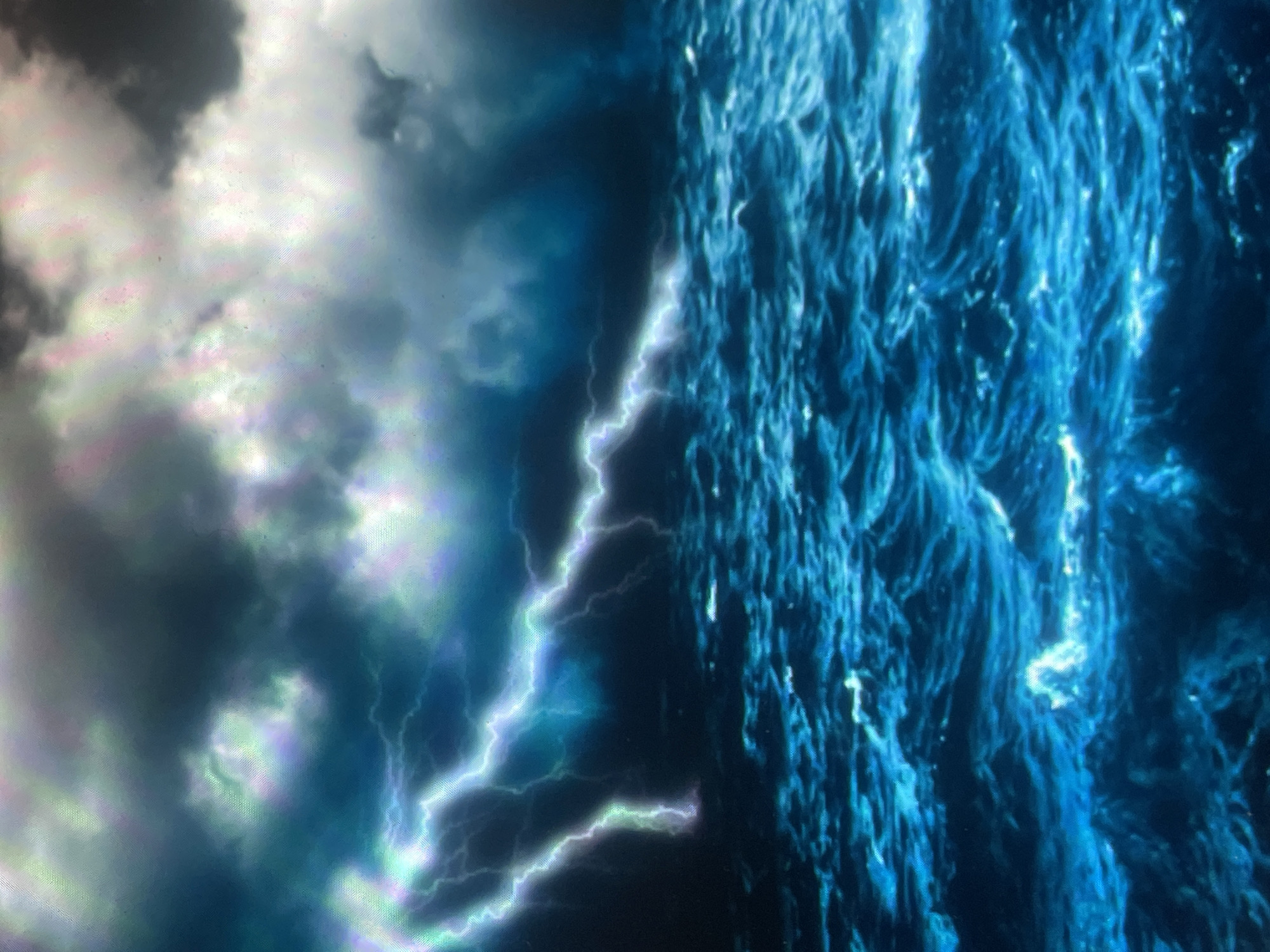 The image displays a violent ocean storm under a dark, turbulent sky. The upper portion of the frame is filled with heavy, dark grey storm clouds, interspersed with luminous, iridescent patches of green, purple, and white light, suggesting atmospheric electrical activity or intense illumination from within the clouds. Multiple streaks of bright, electric blue and white lightning cut across the mid-ground, with prominent bolts visible on the left side extending downwards and a long, horizontal strike stretching across the central frame. The lower half of the image depicts a tumultuous ocean, characterized by deep blue-green waters, significant wave action, and numerous whitecaps, indicating rough seas. No human subjects, man-made objects, or vessels are visible. The scene exclusively presents natural elements of an open sea environment during a storm; consequently, no specific terrestrial location, including the city of Zelenyi Hai, Ukraine, or any urban features, can be identified or inferred from the visual information provided.