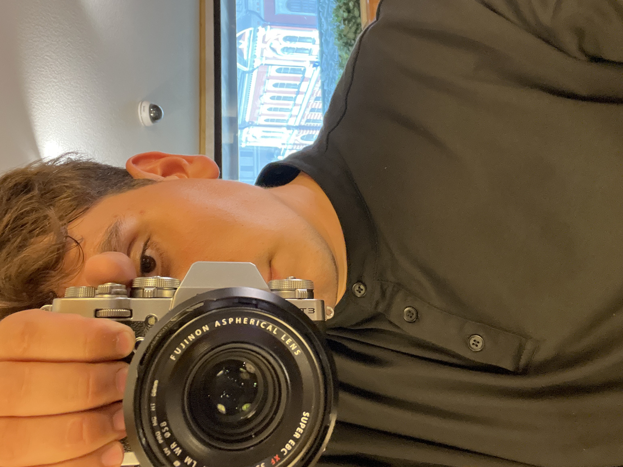 A man is taking a selfie with a digital camera. The camera is silver and has the branding "FUJINON ASPHERICAL LENS" and "SUPER EBC XF" visible on the lens. The man is wearing a black collared shirt with two buttons. Behind him, through a window, is a view of a red building with arched windows, which appears to be a historical structure. In the background, a white security camera is mounted on a ceiling. This building is likely the National Museum of the Republic of Tatarstan located in Kazan, Russia, given its architectural style and historical significance in the city.