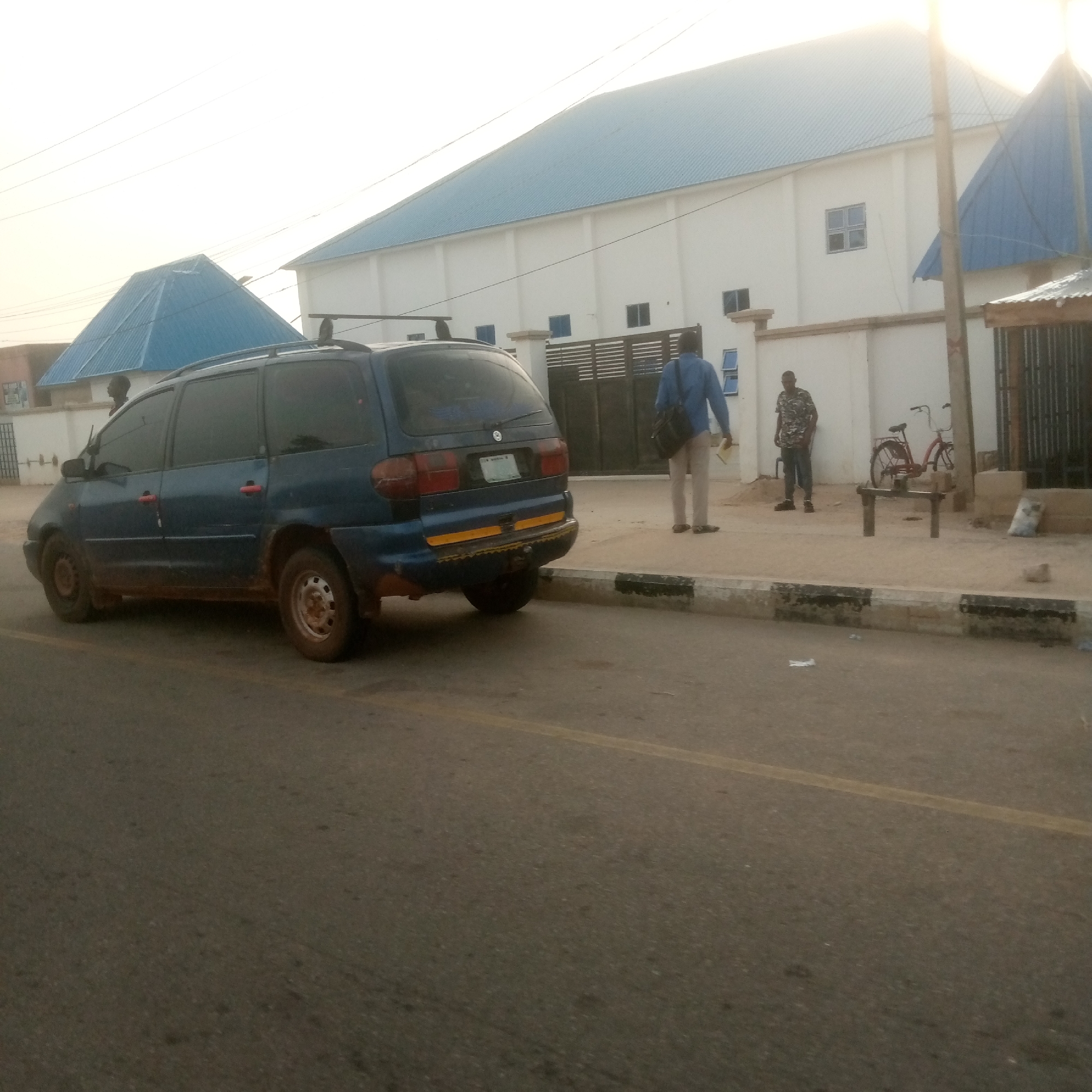 An exterior, ground-level, medium shot shows a blue minivan parked on the right side of a road. The minivan is facing left, and its rear is toward the camera. A man in a blue shirt and light-colored pants is standing on the sidewalk in front of a white building. He is facing away from the camera, and he is holding a yellow item. Another man in a patterned shirt and dark pants is standing to his left, also facing away from the camera. A red bicycle is propped on a stand to the right of the second man. The building behind them has a light blue, sloped roof, and windows are visible on its facade. The sky is overcast.