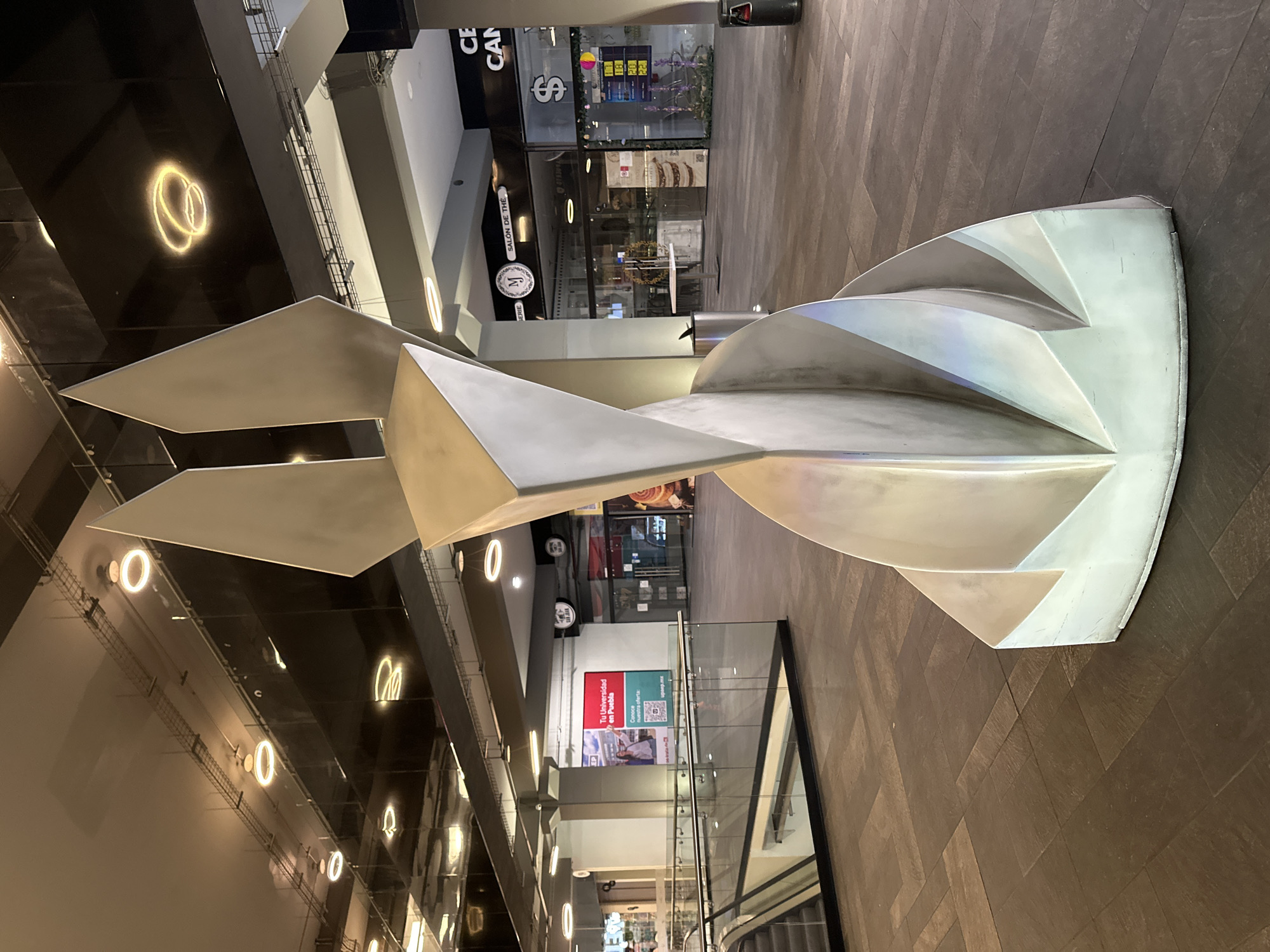 The image displays the interior of a modern commercial complex or shopping mall in Puebla, Mexico, identified by a visible "Tu Universidad en Puebla" (UPAEP) advertisement banner.
A large, abstract sculpture, constructed from reflective, silvery-gray metallic planes, occupies the center foreground. The sculpture features sharp angles, geometric folds, and an upward-tapering design, culminating in two distinct pointed elements. Its base expands in a fanned, layered manner.
The flooring consists of dark gray and brown rectangular tiles arranged in a patterned layout. The ceiling is dark, possibly black, and is equipped with multiple circular, illuminated ring lights that create reflections on the ceiling surface. Exposed overhead conduits are also visible.
To the left, a downward-moving escalator is present, bordered by a clear glass and metal handrail. In the background, a corridor lined with various storefronts is visible. One storefront features a sign reading "SALON DE THÉ" with an "MJ" logo. Next to it, another establishment displays a prominent dollar symbol ('$') and a digital screen showing numerical values such as "1.1," "1.8," "2.0," and "2.2," indicative of currency exchange rates. Further down the corridor, signs displaying "SAN JUAN" are visible. The aforementioned "Tu Universidad en Puebla" banner, featuring the UPAEP logo and "upaep.mx," is prominently displayed on a wall.
Two silver-colored, cylindrical trash receptacles are positioned within the scene: one to the immediate right of the central sculpture and another further back near the "SALON DE THÉ" store. The area is artificially lit, and no individuals are present within the frame.