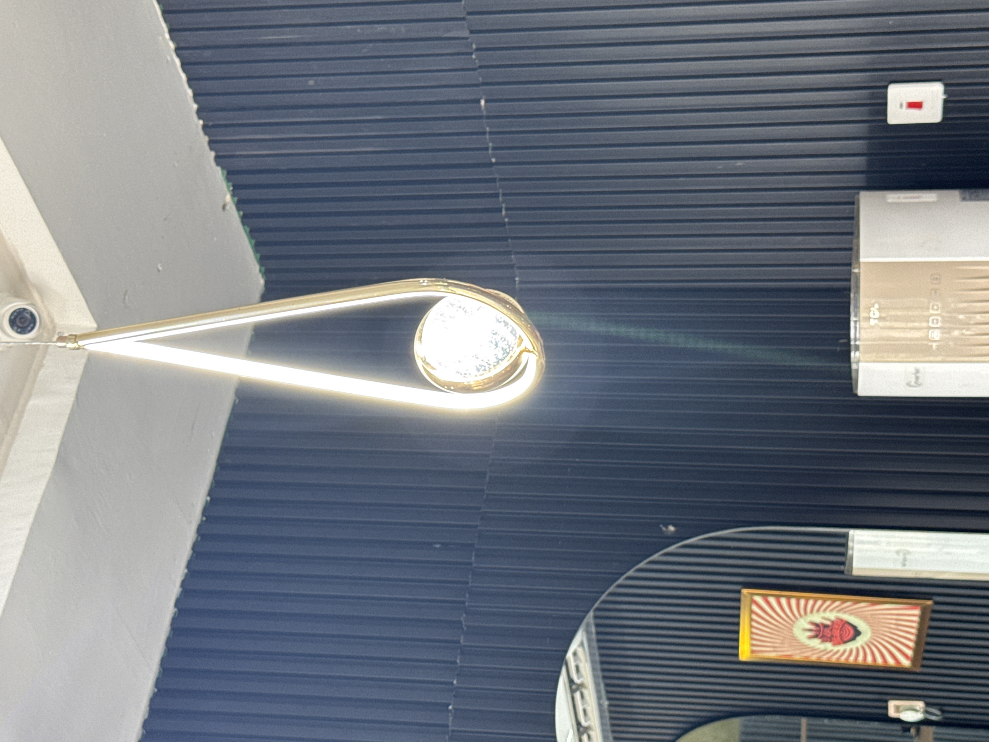The media file captures an upward view of an indoor space, likely a modern commercial establishment in Kurunduma, Nigeria. The dominant features include a striking, golden, loop-shaped light fixture with bright internal LEDs, suspended from the ceiling. Most of the background consists of a dark, horizontally slatted or corrugated ceiling.
On the far right side of the ceiling, a silver-colored air conditioner unit is mounted, with a small white box containing a red indicator (possibly a fire alarm or emergency button) positioned above it. To the left, a light-colored, possibly off-white, wall or structural beam intersects with the dark ceiling. A small, white security camera with a dark lens is affixed to this wall in the lower-left corner of the frame.
In the bottom right, a curved, reflective surface, likely a mirror, shows a partial reflection of the room. Within this reflection, or possibly mounted on an adjacent surface, a small framed artwork is visible, featuring a red, stylized fish-like symbol against a background of radiating red and white stripes.
No people are present, and no specific activity is taking place. The scene appears static, showcasing the architecture and interior design. The bright artificial light from the fixture creates a subtle lens flare across the image. The text "TCL" is clearly visible on the air conditioner unit, along with some less distinct numerical markings (e.g., "180000").
