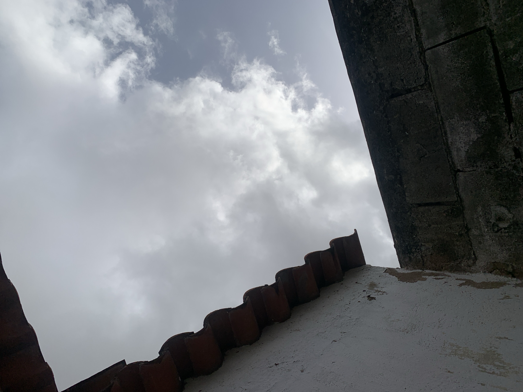 The image captures an upward-angled view of a cloudy sky framed by architectural details, set in Santa Ana, Venezuela. The upper and central portions of the frame are dominated by an expansive sky filled with a mix of white and gray clouds, suggesting an overcast or partly cloudy daytime. Lighter patches of blue-gray sky are intermittently visible between the cloud formations.
On the lower left, dark reddish-brown, traditional corrugated roof tiles ascend diagonally from the corner, showcasing their distinctive curved form. Adjacent to and below these tiles, towards the bottom right, is a textured, off-white wall. This wall displays signs of age and wear, with areas of peeling paint or rough plaster.
Along the entire right side of the image, a large, dark, and robust structure rises vertically. This appears to be a weathered stone or concrete wall, composed of discernible blocks and exhibiting a coarse, aged texture, possibly indicative of a historic building or fortification. The perspective is from a low vantage point, looking upwards towards the sky and the convergence of these architectural elements. No human figures or activities are depicted in the static scene. There is no visible text.