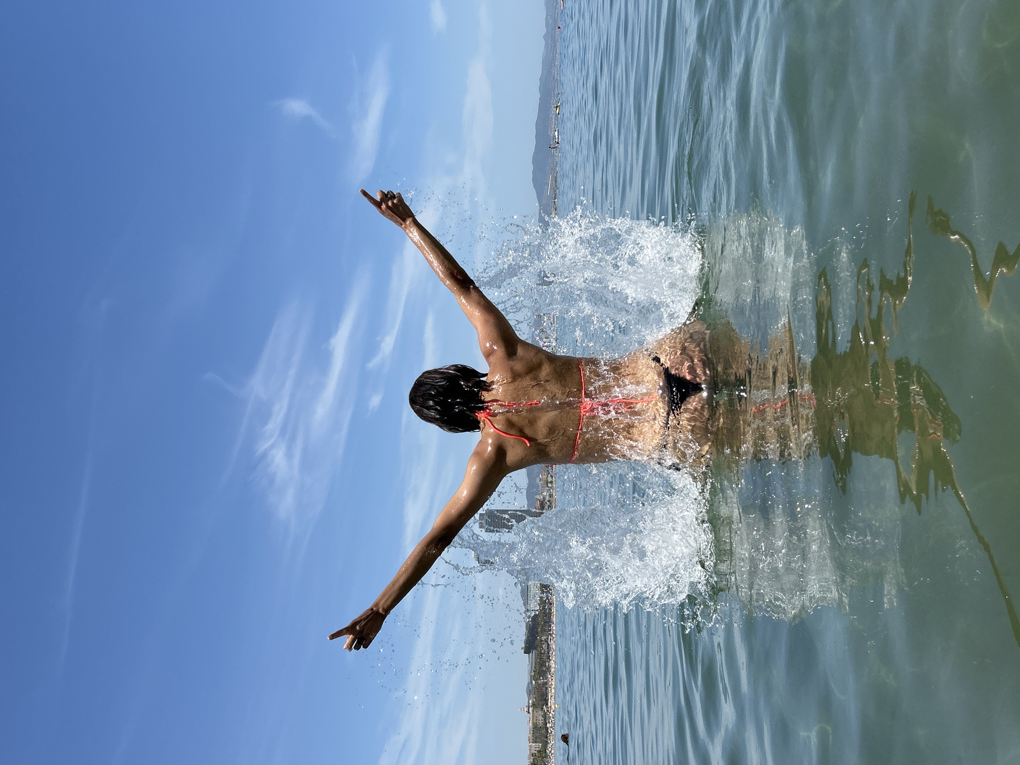 A woman in a black bikini is emerging from the water, creating a large splash. Her arms are raised and extended outwards, and her hands are pointed upwards. The background shows a clear blue sky with wispy clouds, and a city skyline with buildings and a beach. The water is a clear blue-green, with sunlight reflecting on the surface. The location appears to be the Mediterranean Sea, off the coast of Barcelona, Spain.
