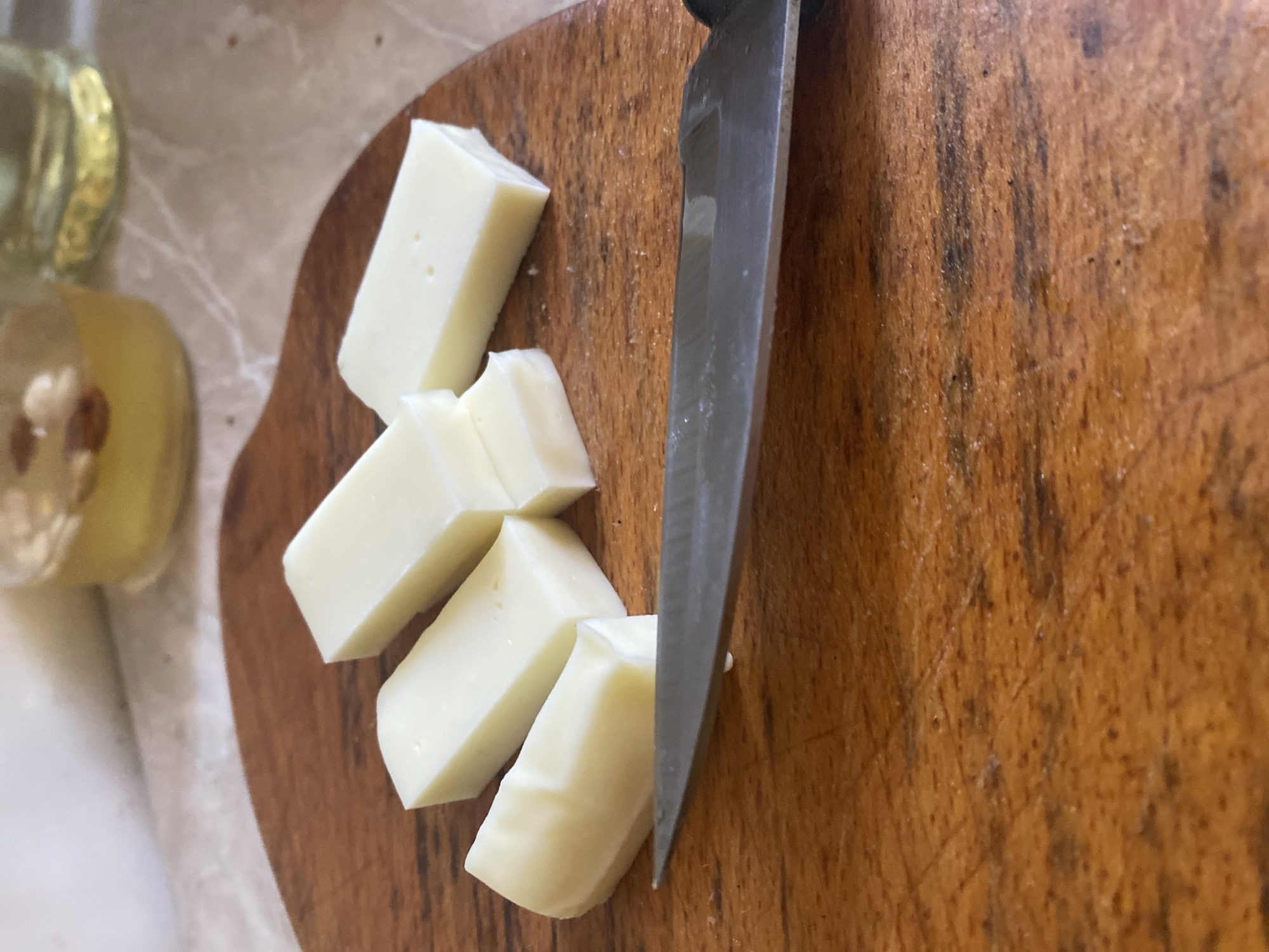 The image shows a close-up, overhead view of a wooden cutting board with several pieces of white cheese. A silver knife is positioned on the cutting board, its blade angled towards the cheese. The cheese has been cut into roughly uniform rectangular pieces, some of which are slightly rounded. The cutting board has a visible wood grain pattern and appears to be well-used, with some scratches and darker marks. To the left of the cutting board, out of focus, is a glass bottle with a yellowish liquid, possibly oil, and a white cloth or napkin. The lighting suggests it could be daytime, but no specific time is evident. The scene appears to be a simple preparation of food, likely in a kitchen or similar setting. There is no visible text in the image.