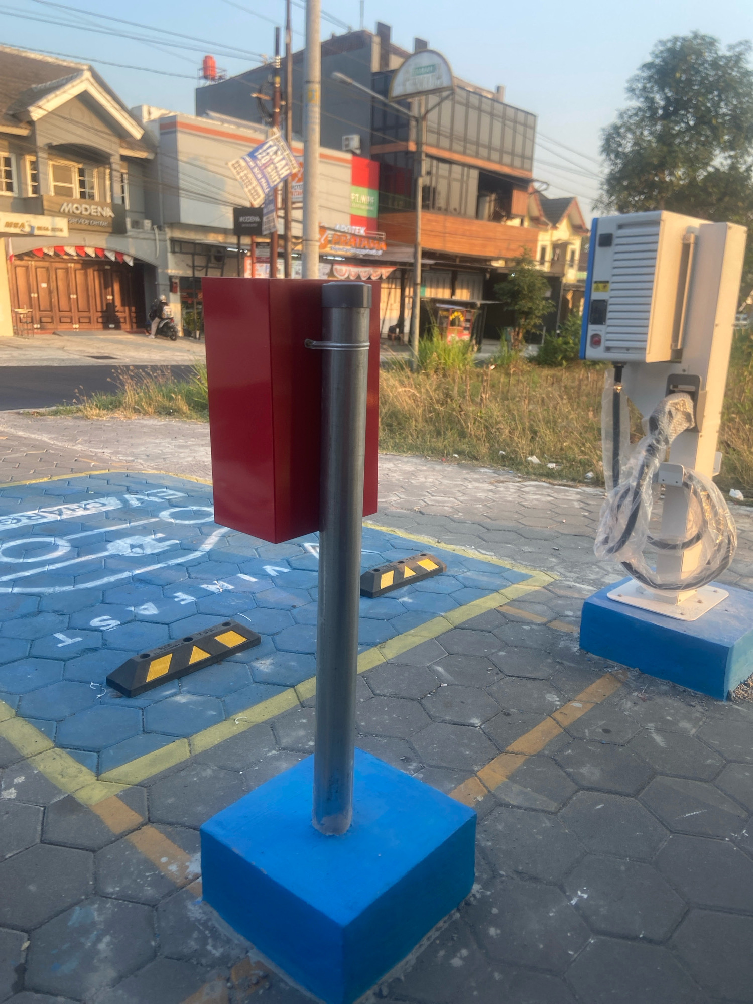 The image depicts an outdoor scene in the city of Sleman, Indonesia, featuring what appears to be an electric vehicle charging station. In the foreground, a red box mounted on a metal pole stands on a blue-painted hexagonal pavement. Beside it, a white charging unit with a coiled cable covered in plastic wrap is also situated on a blue base. Yellow lines mark the pavement, and two black and yellow chocks are placed on the ground. In the background, buildings line a street, including a structure with "MODENA service center" signage. A person on a motorcycle is visible near the buildings. The environment suggests a commercial area with some vegetation and overhead utility lines. The lighting indicates it is either early morning or late afternoon.