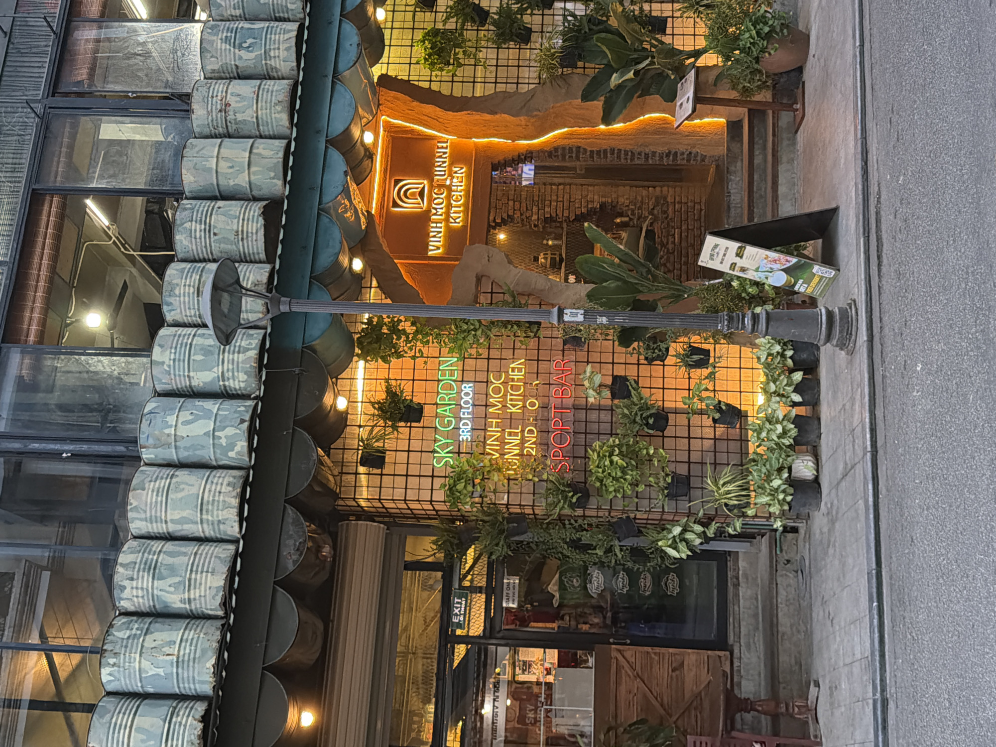 The image shows the exterior of a restaurant and bar in Thuận Hóa Ward, Vietnam. The building has a dark facade with large windows on the upper floor. Below the windows, several large, cylindrical metal barrels with a camouflage pattern are stacked and arranged along the roofline.
The ground floor features a prominent entrance to "Vinh Moc Tunnel Kitchen" with a glowing orange neon sign. The entrance is framed by a rough, textured material that resembles a tunnel opening, also illuminated with orange light. To the left of the entrance, a neon sign reads "SKY GARDEN 3RD FLOOR," "VINH MOC TUNNEL KITCHEN 2ND FLOOR," and "SPORT BAR." This area is decorated with numerous potted plants, hanging vines, and a wire mesh grid on the wall.
A street lamp stands in front of the entrance. An A-frame sign with a "GRAND OPENING" message and a QR code is placed on the sidewalk. The sidewalk is paved with gray concrete tiles, and a street runs in front of the establishment. The scene is illuminated by the neon signs and overhead lights, suggesting it is evening or nighttime. No people are visible in the image.