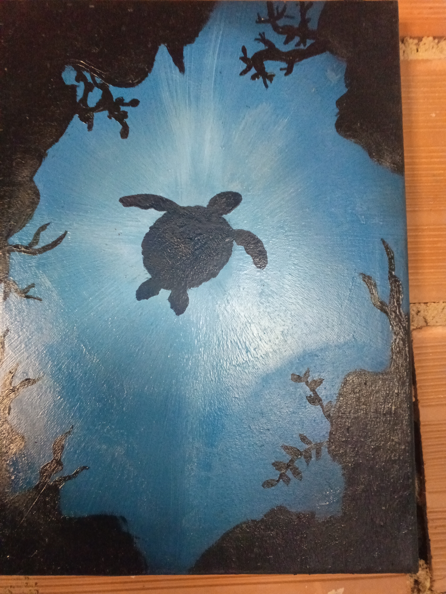 The image is a painting depicting a sea turtle swimming upwards in an underwater cave environment. The turtle is dark in color and centrally positioned. Light rays emanate from the top of the painting, suggesting sunlight penetrating the water. The cave walls are dark and textured, with silhouettes of plant-like structures or coral formations extending from them. The background is a gradient of blue, lighter near the top. The painting appears to be resting against a wooden wall or surface. There is no information about the city of Tinaquillo, Venezuela.
