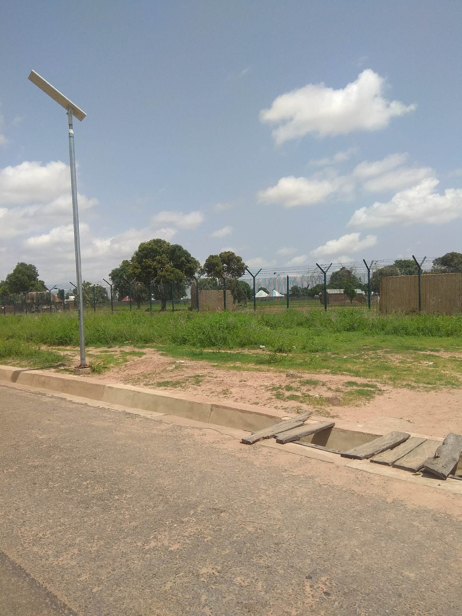 The image depicts an outdoor scene in Jalingo, Nigeria, likely in a semi-urban or industrial area. In the foreground, a paved road with a gravelly texture is visible, bordered by a concrete curb. Adjacent to the curb is a drainage ditch with several wooden planks laid across it, suggesting a makeshift crossing or repair.
A solar-powered street lamp stands on the left side of the image, its metallic pole extending upwards towards a partly cloudy blue sky. The background features a strip of green grass and vegetation, behind which a chain-link fence with barbed wire is erected, indicating a boundary or security measure. Beyond the fence, several trees with lush green foliage are scattered, and some buildings with light-colored walls and darker roofs can be seen. The overall environment suggests a sunny day with moderate cloud cover. No people or animals are present in the frame.