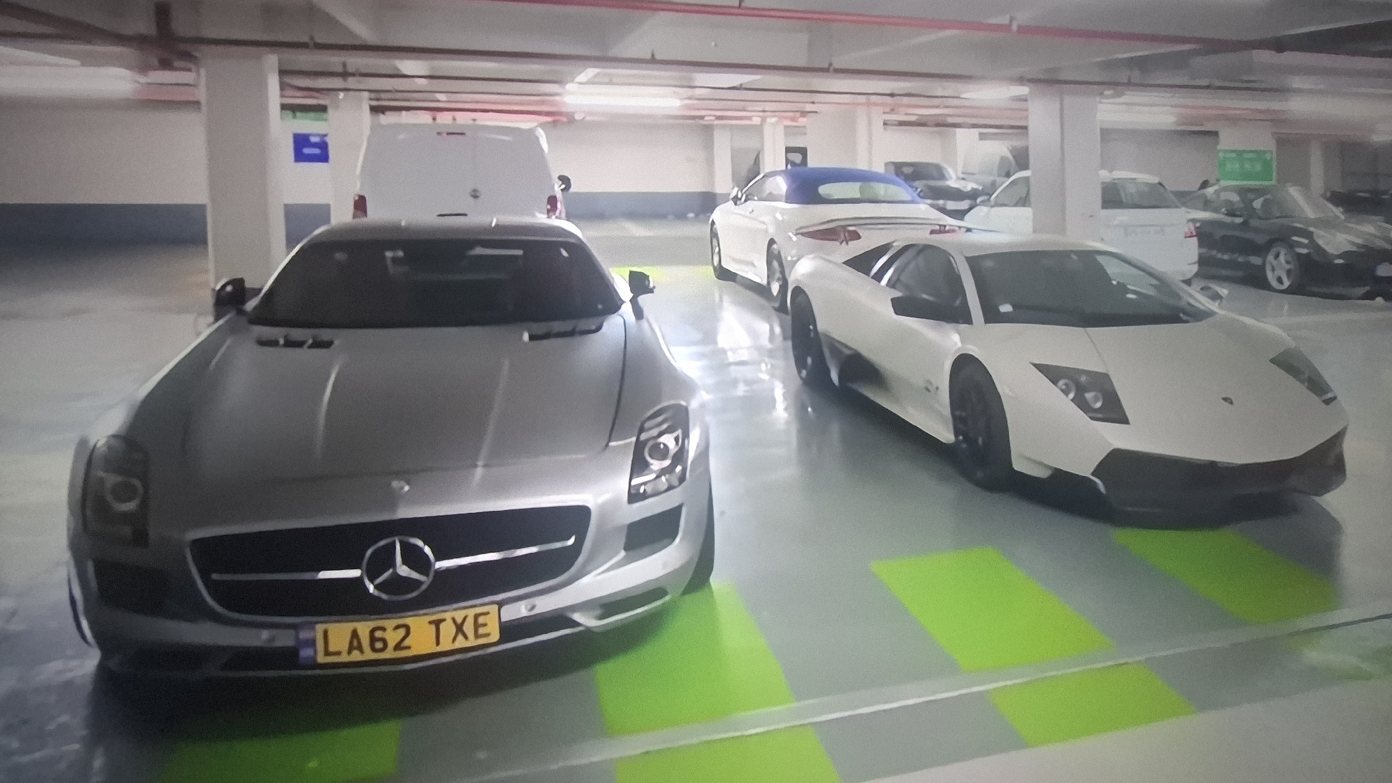The image depicts an underground parking garage in an unknown city, with a silver Mercedes-Benz SLS AMG (license plate LA62 TXE) parked in the foreground. To the right of the Mercedes is a white Lamborghini Murciélago. Behind these vehicles, there is a white convertible, a white van, and several other obscured cars. The parking space lines are painted with alternating green and gray stripes. Overhead lighting is visible in the ceiling. There is a sign with a blue background hanging from one of the pillars.