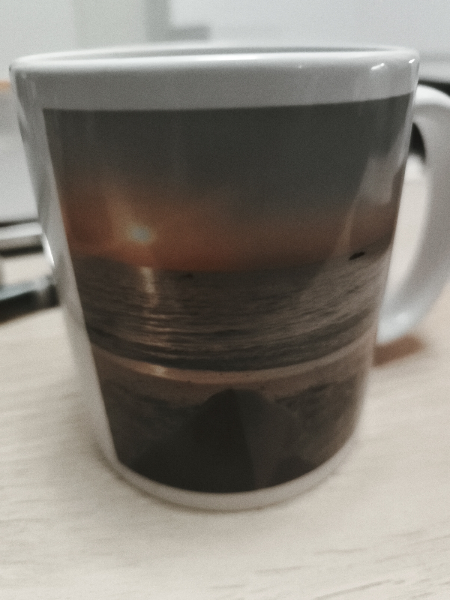 A white ceramic mug with a handle on the right is centrally positioned, resting on a light-colored, possibly wooden or laminate, surface. The mug features a panoramic print of a tranquil seascape at either sunset or sunrise. A distinct, bright sun is visible low on the horizon, casting a warm, orange-yellow light across the sky and reflecting on the gently rippling surface of the ocean. A dark, indistinct landmass or beach appears in the foreground of the image printed on the mug. The overall lighting of the photograph itself is soft, indicating an indoor setting. The background outside of the mug is out of focus, showing blurry, light-toned elements consistent with an interior room or office environment. No people or visible text are present in the image. The scene depicted on the mug evokes a sense of natural beauty and calm. This media file was captured in Huntingdonshire, United Kingdom.