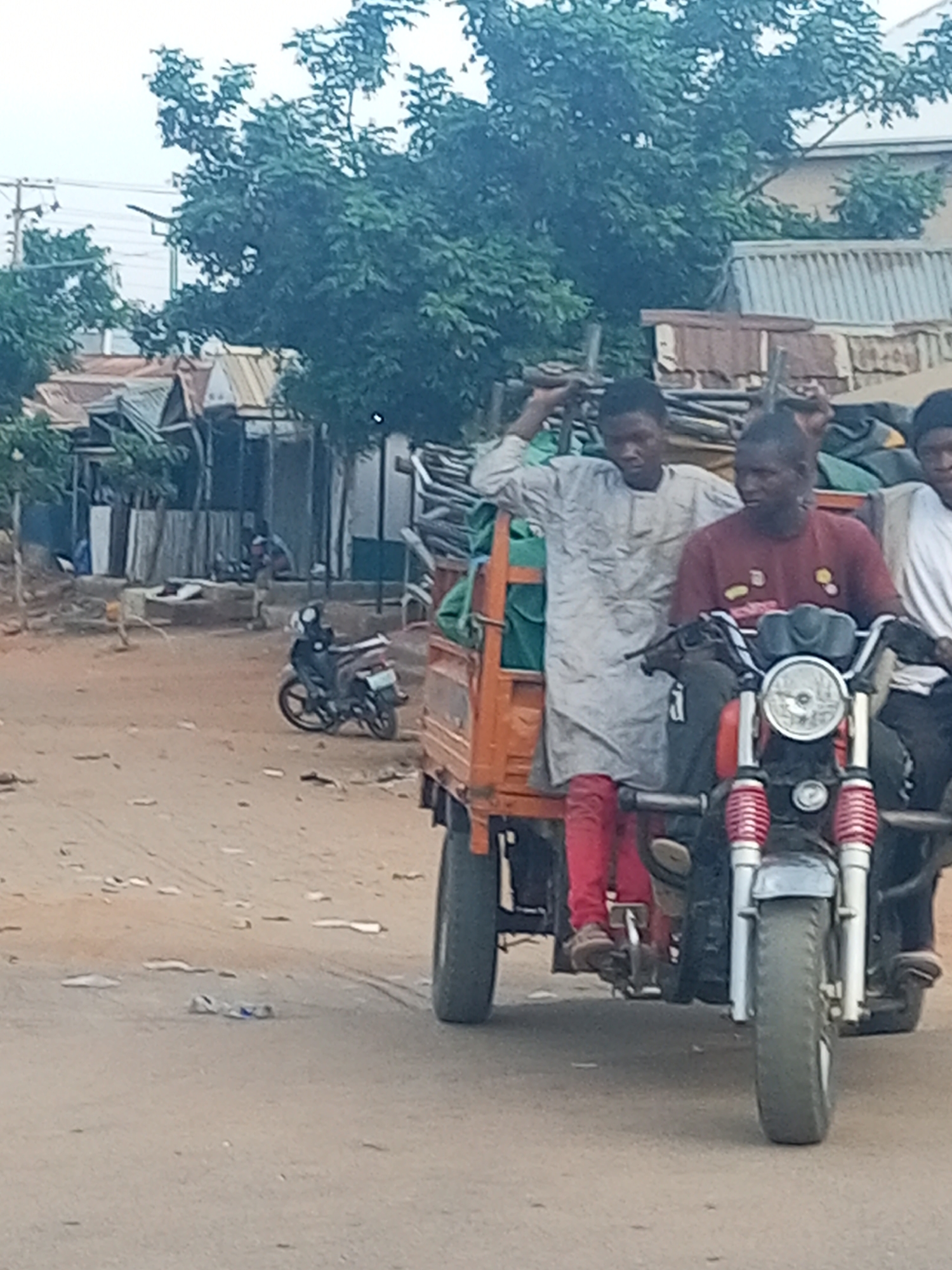 The image depicts a street scene in what appears to be Tempure, Nigeria. Three men are riding on a three-wheeled motorcycle-rickshaw. The vehicle is orange and has cargo in the back. The houses in the background are mostly shacks constructed of various materials. Another motorcycle is parked on the side of the street. The road is dirt and scattered with trash. Trees are visible in the background.