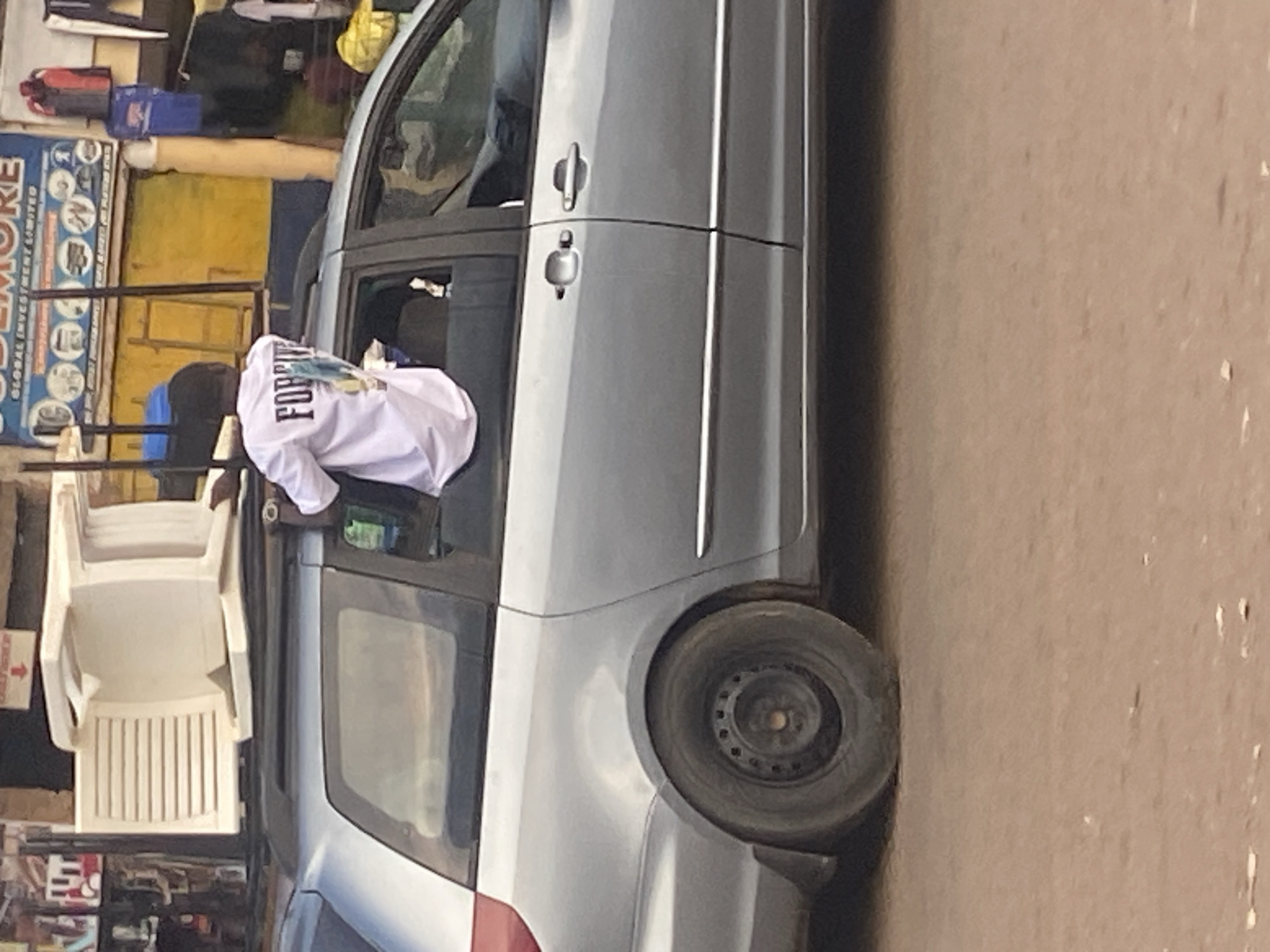 A young male wearing a white t-shirt with "FOREVER" printed on the back is leaning out of the open passenger window of a silver minivan. He is sitting with his legs inside the vehicle and his upper body outside, with his left arm resting on the roof rack. The minivan's rear tire is flat. The vehicle is parked on a dirt road in front of a building with a yellow wall and a blue sign that reads "BUKURE GLOBAL INVESTMENT LIMITED." There are items displayed for sale in front of the building, including clothing and what appears to be plastic furniture stacked on the roof of another vehicle. The scene is consistent with a street market in Bukuru, Nigeria.