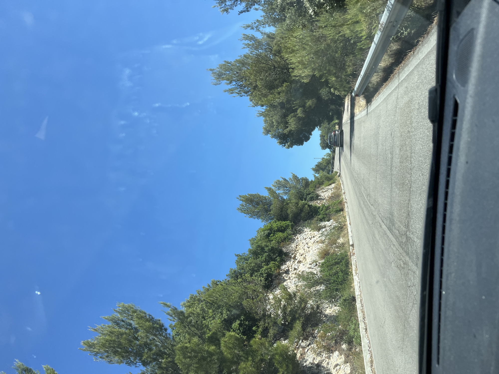 The image shows a paved road winding uphill through a landscape with rocky terrain and lush green trees on the left. The sky is a clear, bright blue with a few wispy white clouds. A silver SUV is visible driving ahead on the road. The perspective suggests the photo was taken from the driver's seat of a vehicle. The scene is characteristic of a scenic route in a Mediterranean coastal area, possibly within the Gargano National Park near Mattinata, Italy, known for its rugged coastline and abundant Mediterranean vegetation. The overall impression is one of a sunny day on a road trip through a natural, undeveloped environment.