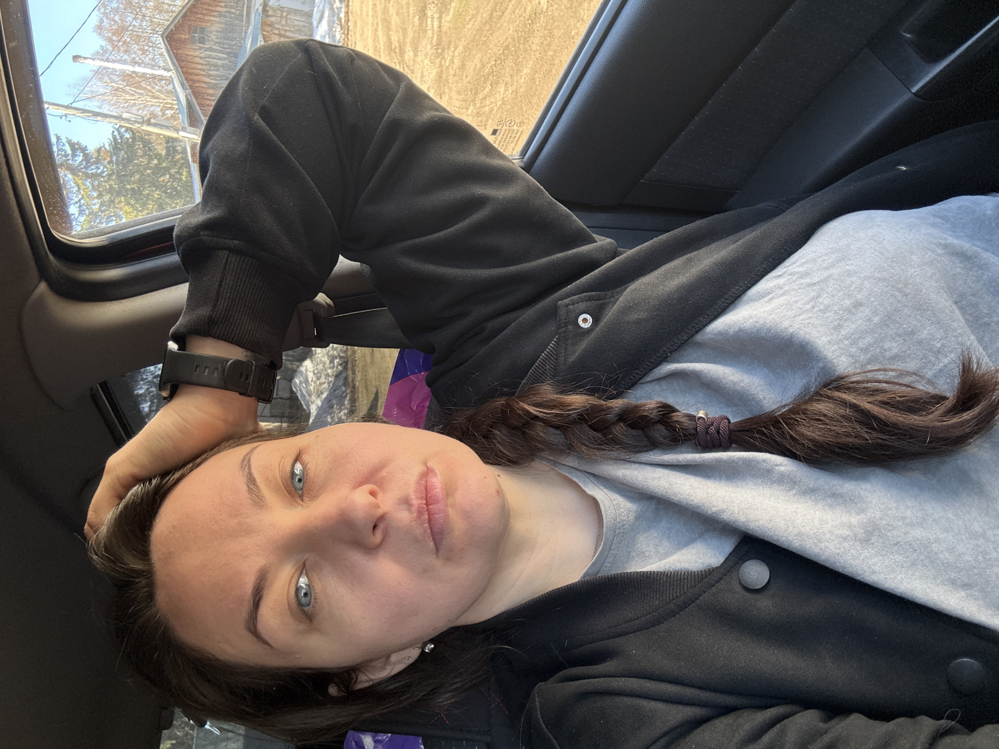 A portrait-style selfie taken inside a vehicle features a person, likely female, with medium-dark brown hair styled in a braid secured with a dark hair tie. She has prominent blue eyes and a neutral expression, looking directly at the camera. She wears a light grey long-sleeved shirt and a black jacket with snap buttons, and a black digital watch is visible on her left wrist.
The background outside the vehicle's window reveals a clear blue sky, bare trees, and a rustic wooden building with a visible chimney, suggesting an early spring or late autumn setting in a rural or suburban area. The ground outside appears dry and light-colored. The bright, natural lighting indicates it is daytime. The person appears to be seated or leaning back in the car. No visible text is clearly discernible in the image. The scene is consistent with a location such as Chaikovsky, Russia.