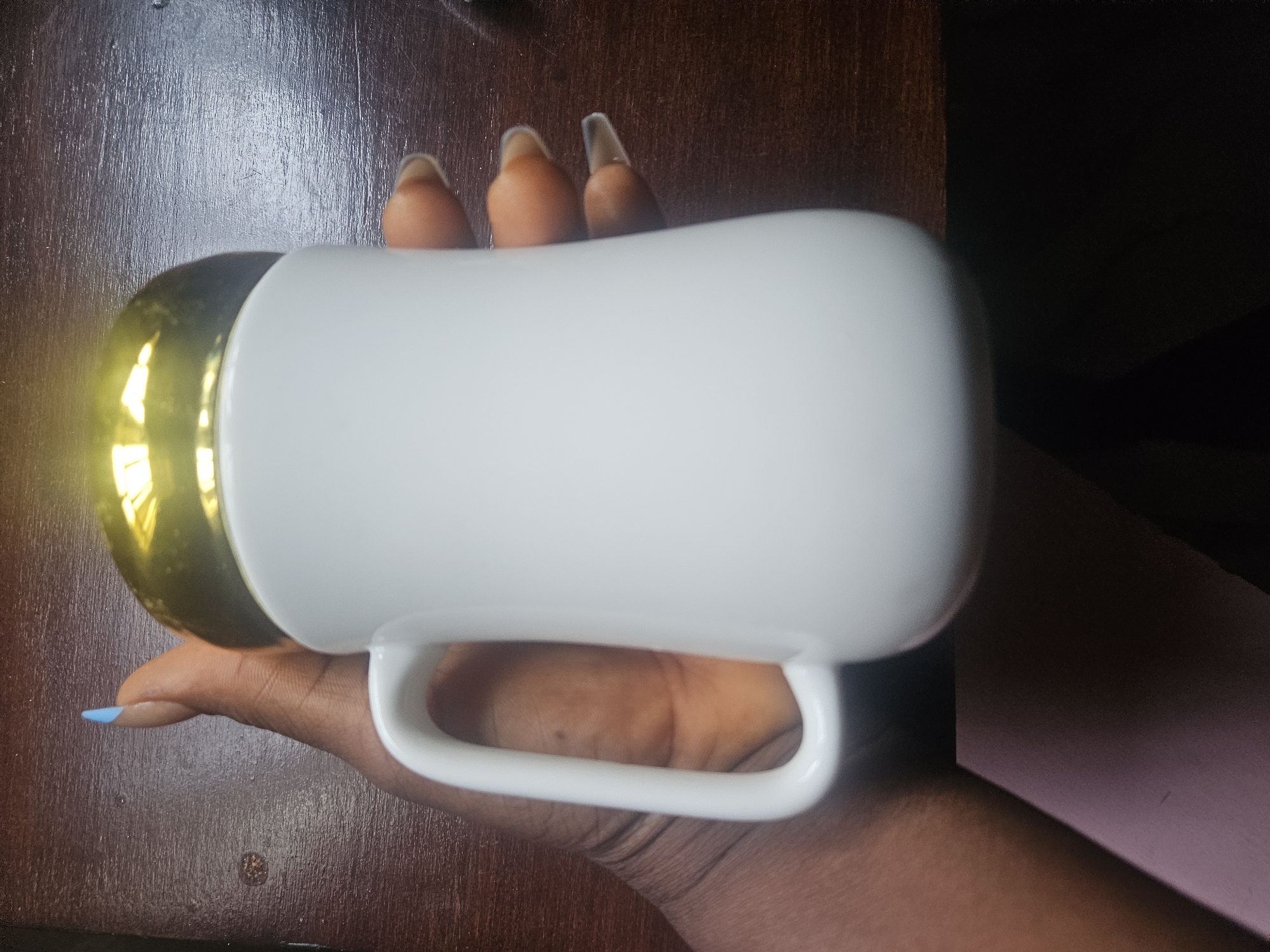 A person's hand, with light blue painted fingernails on the index finger and natural-colored, long, pointed fingernails on the other fingers, holds a white ceramic mug with a gold lid. The mug has a rounded bottom and a C-shaped handle. The hand and mug are positioned over a dark wooden surface. The scene is located in the city of Masaka, Nigeria.
