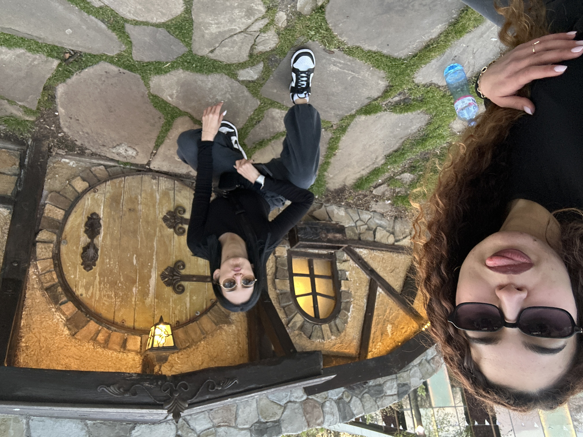 An outdoor, vertically oriented photograph, taken from a slightly elevated perspective, captures two young women posing in front of a whimsical, fairy-tale-like building. The image is inverted relative to standard viewing.
In the foreground, on the right side of the frame (when viewed correctly), a woman with long, curly brown hair and dark sunglasses playfully sticks out her tongue. She wears a black top, and her right hand, adorned with a gold bracelet and ring, shows light-colored nail polish. A blue plastic water bottle with "DEVIN" visible on its label lies on the flagstone path near her.
To the left and slightly behind her, the second woman is seated on the ground with her legs bent. She wears a black long-sleeved top, dark grey sweatpants, and black and white sneakers. Dark sunglasses cover her eyes, and a smartwatch is visible on her left wrist. She looks towards the camera with a slight pout.
The background features a charming structure with a large, round wooden door and a smaller arched window, suggesting a Hobbit-house or fantasy cottage design. The building has textured, golden-brown walls framed by dark wooden beams, and a stone foundation. A small, illuminated lantern hangs near the circular door. The ground is a flagstone path with grass growing between the stones. The lighting suggests it is daytime, possibly overcast or late afternoon. The overall mood is casual and playful, set in what appears to be a unique outdoor location in Burgas, Bulgaria.