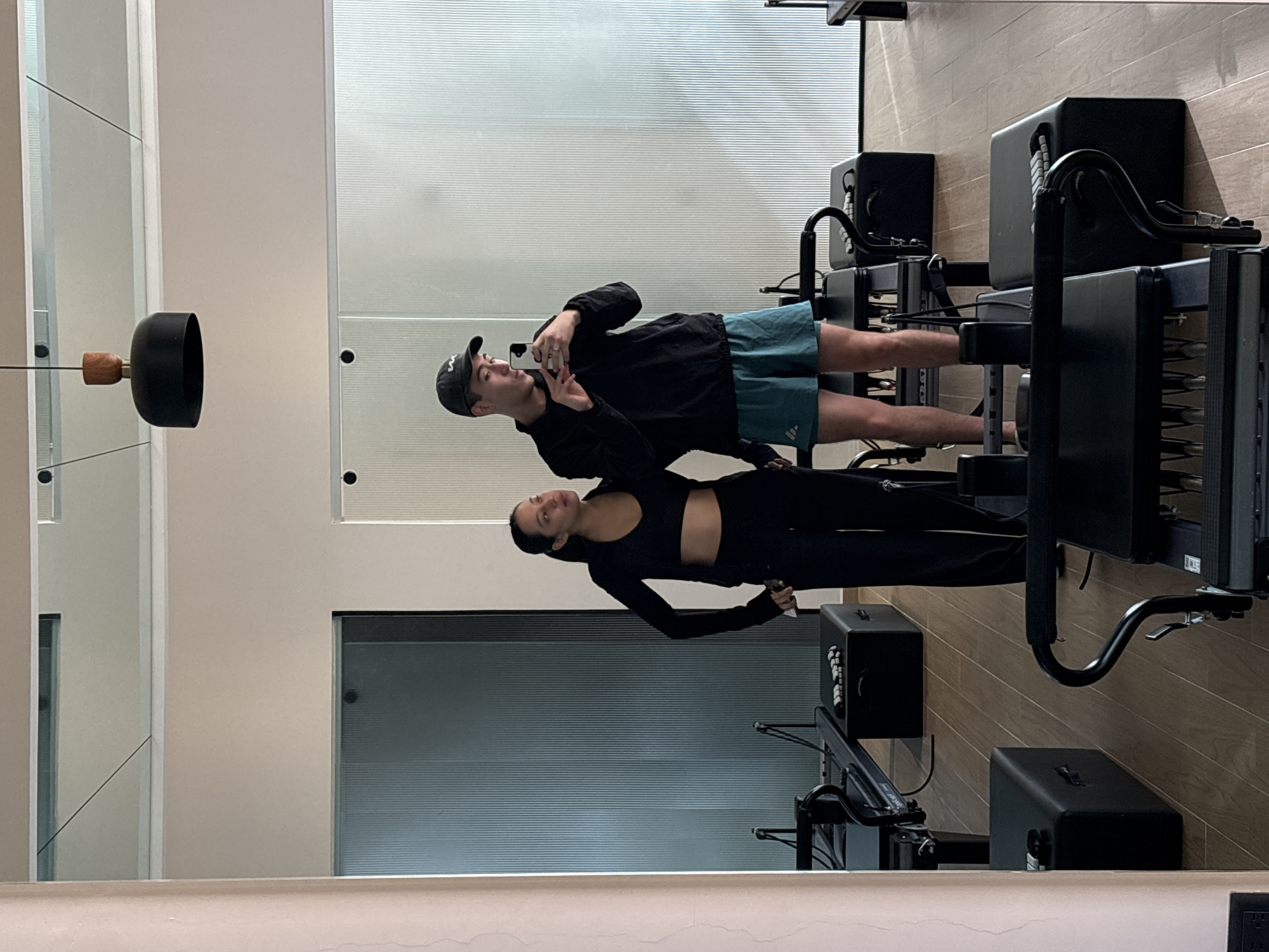 The image captures an indoor scene, likely a modern fitness studio in San Bernardino Tlaxcalancingo, Mexico, through a reflection in a large mirror. The primary subjects are two individuals: a man and a woman.
The man, positioned on the left side of the reflection, wears a black baseball cap, a black long-sleeved athletic top, and teal-colored shorts featuring a white "Adidas" logo on the left leg. He is holding a smartphone to take a mirror selfie, and his left hand is raised, forming a peace sign. His gaze is directed towards the phone screen.
Standing to the right of the man in the reflection is a woman, dressed in a black athletic crop top or sports bra and black leggings. Her dark hair is pulled back, and her expression is neutral as she looks towards the mirror or phone.
The background of the reflection reveals a clean, minimalist setting. Two black Pilates reformer machines are visible, suggesting the studio's purpose. The floor appears to be made of light-colored wood. The walls are a neutral, light beige, with sections that include horizontally textured glass or blinds, possibly part of windows or room dividers. A distinct black, modern light fixture with a wooden accent is suspended in the upper left of the reflection. The overall ambiance is bright and contemporary.