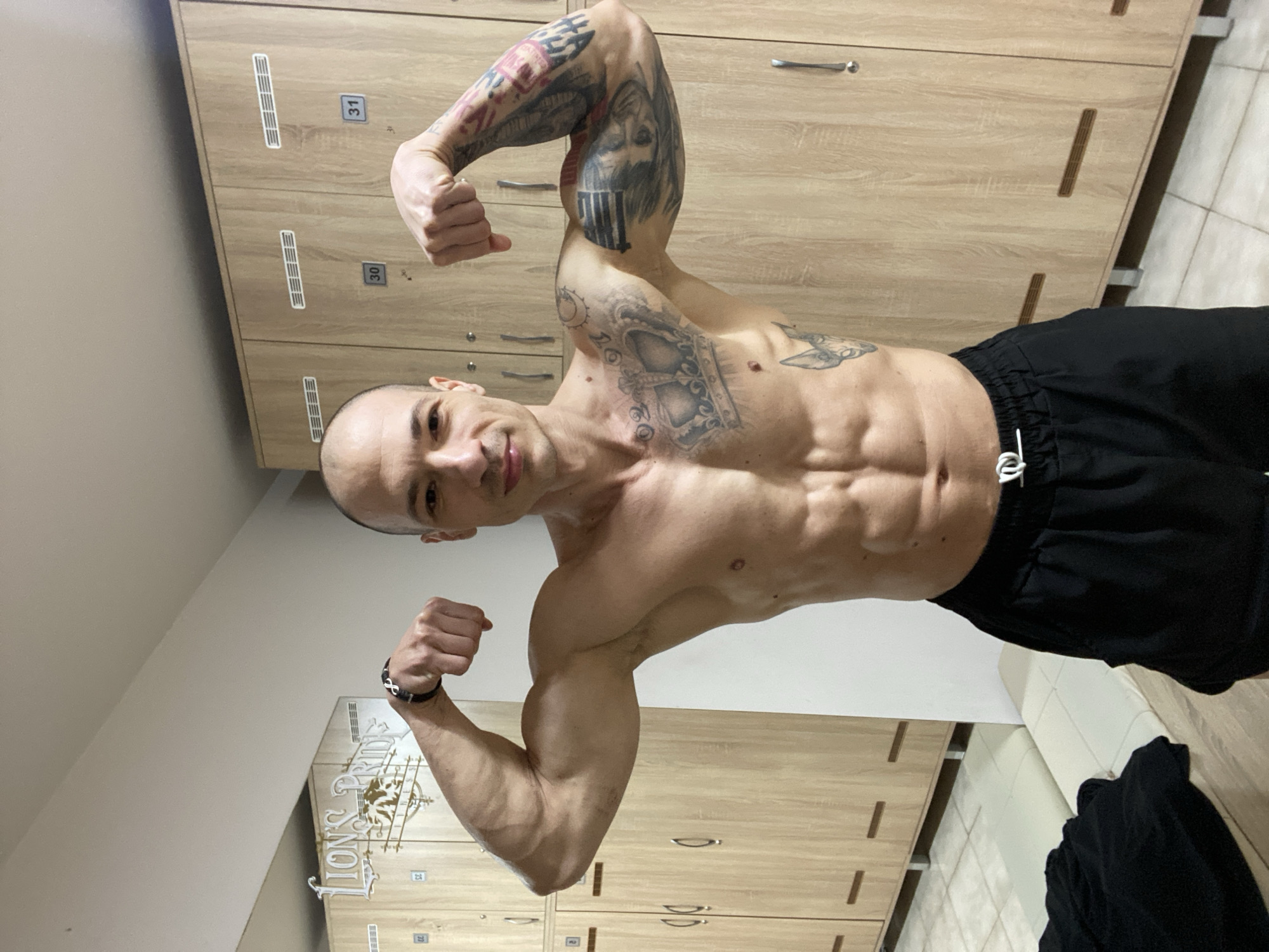 A bald man is in a room with lockers, flexing his biceps. He has tattoos on his arms, chest, and stomach. He is wearing black shorts. The lockers are light brown wood. The scene appears to be inside a gymnasium or changing room. There is no other information to suggest the location is in Pleven, Bulgaria.