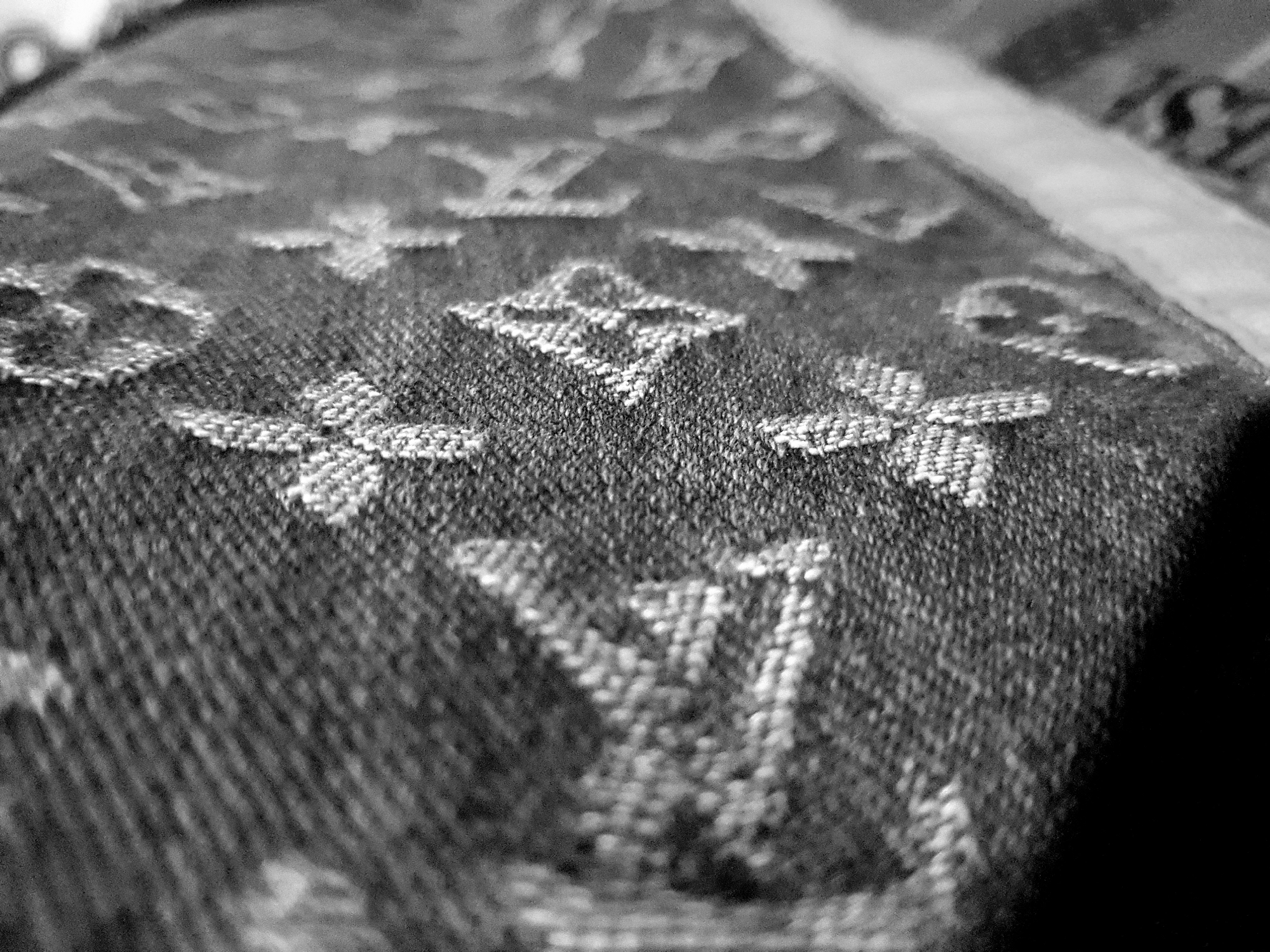 A black and white close-up image shows a textured fabric with a repeating pattern of stylized flowers and diamonds. The fabric appears to be woven, with visible threads creating a raised pattern. The lighting creates shadows and highlights, emphasizing the texture and depth of the fabric. The composition is abstract, focusing on the intricate details of the material. The image is not taken in any specific city; it is a detail shot of a fabric.