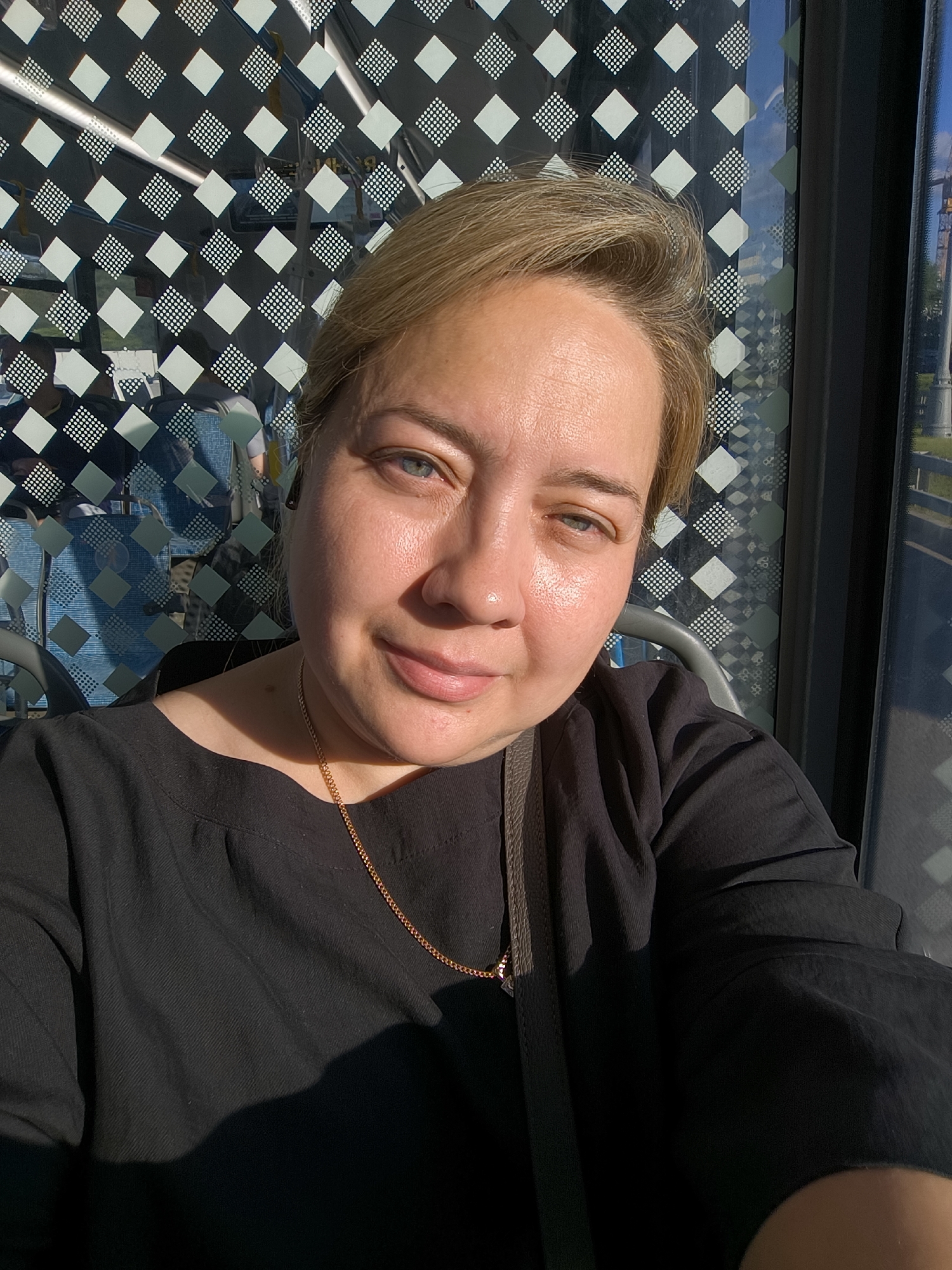 A woman with blonde hair and blue eyes is taking a selfie. She is wearing a black top and a gold necklace. She is sitting on a bus with a patterned window behind her. The sun is shining through the window, casting a shadow on her.