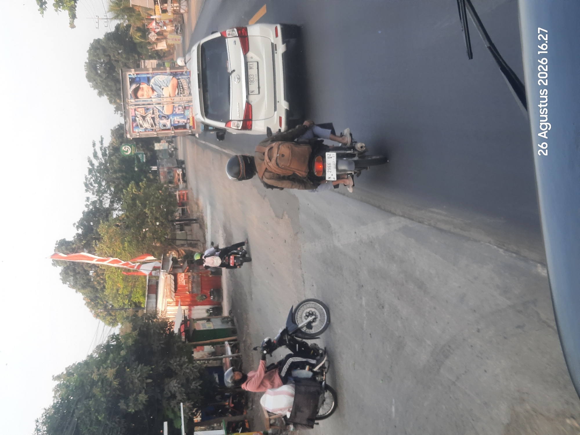 The image captures a street scene in Sukokerto, Indonesia. In the foreground, a black motorcycle with a rider wearing a pink long-sleeved shirt and a helmet is moving to the left, carrying a large white sack in a black carrier. To the right of this motorcycle, another motorcycle with two occupants, both wearing helmets, is also moving to the left. The passenger in the rear is wearing a brown backpack.
Further down the road, a white minivan with license plate "N 1833 MX" is visible. Beyond the minivan, a truck with a colorful advertisement featuring a boy is traveling in the same direction. In the background, there are trees, buildings, and the Indonesian flag displayed on a pole. The road surface appears to be asphalt, with some debris and tire marks. The sky is overcast. The timestamp in the bottom right corner indicates "26 Agustus 2026 16.27".