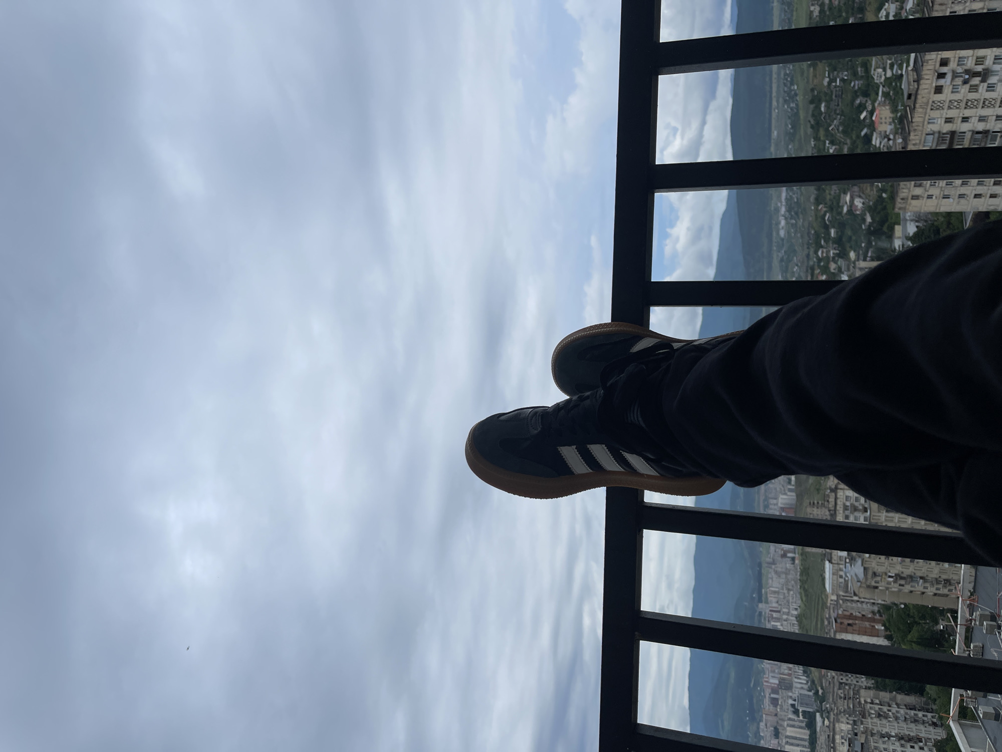 This is a first-person perspective, waist-up shot of a person relaxing on a balcony overlooking a city. The person is wearing black pants and black athletic shoes with white stripes and gum soles. Their legs are crossed, and their feet are resting on a black metal railing. The background reveals a sprawling cityscape with numerous apartment buildings of varying architectural styles and heights, densely packed together. Beyond the city, rolling green hills and mountains are visible under a cloudy, overcast sky. The presence of apartment buildings and the hilly topography strongly suggest the location is Tbilisi, Georgia, a city known for its distinctive Soviet-era architecture and mountainous surroundings. The overall mood is one of quiet observation and relaxation.