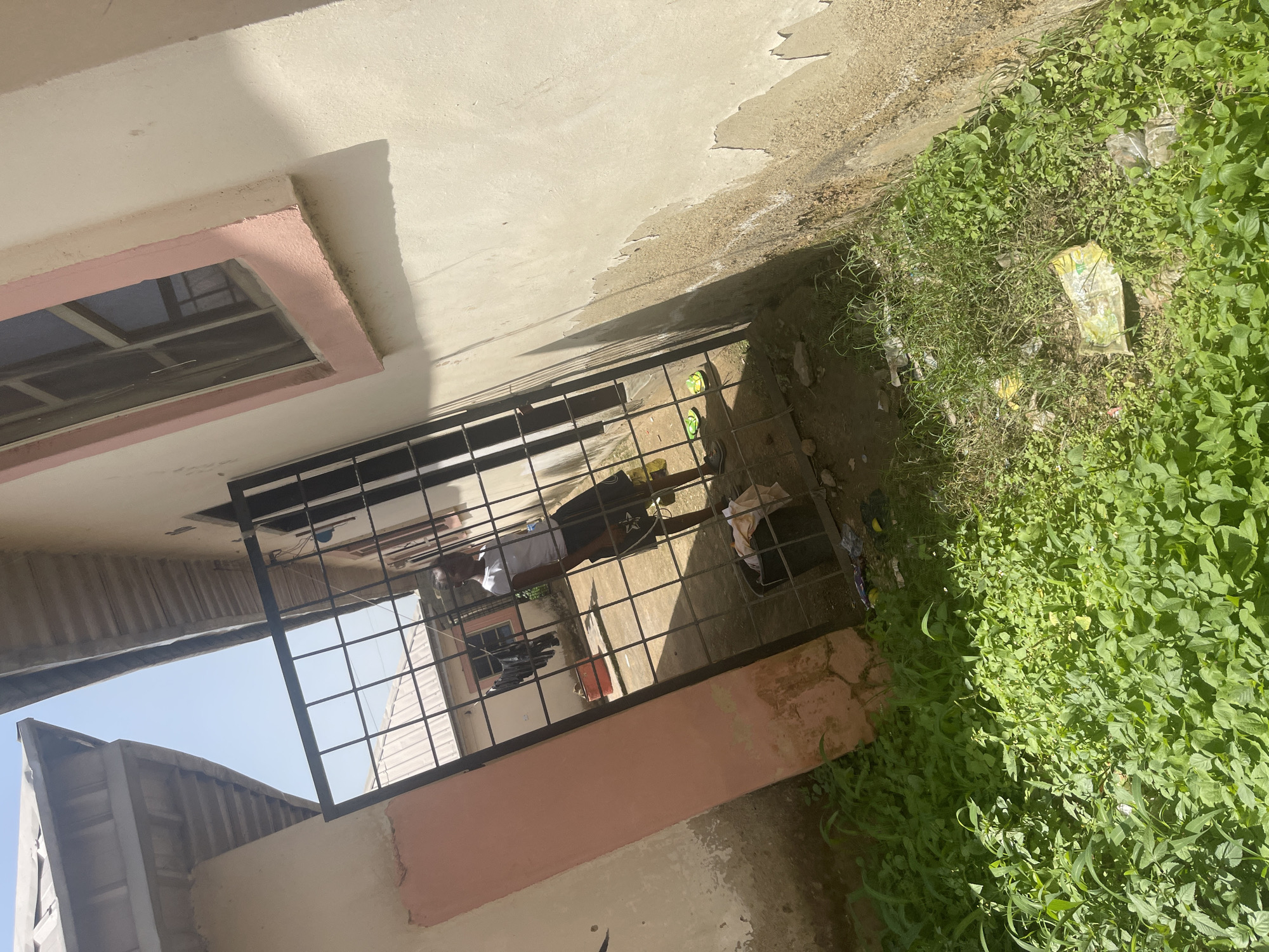 This media file depicts an outdoor scene in Rafin Par, Nigeria. The foreground features lush green vegetation, with some scattered debris, including plastic bags and wrappers, interspersed within the foliage. A concrete pathway, or possibly the side of a building, runs along the edge of the vegetation. Beyond this, a black metal gate or grill is visible, enclosing a courtyard or passage.
Through the gate, a person wearing a white t-shirt and dark shorts can be seen standing. Another person, partially visible, is also present in the same area. The architecture suggests a residential or functional building, with a window framed in pink visible on the left side. The overall impression is one of an outdoor space, possibly a yard or alleyway, adjacent to a building. The sunlight suggests it is daytime, and the presence of vegetation implies a warm climate. There is no visible text in the image. The scene appears to be a casual, everyday setting rather than a specific event.