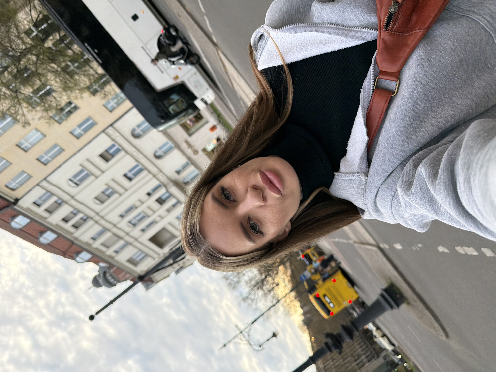 A medium-shot photograph depicts a young woman wearing a grey zippered hoodie, a black turtleneck, and a brown crossbody bag. She is looking directly at the camera with a neutral expression.
The setting appears to be a street in Berlin, Germany. In the background, there is a cream-colored building with multiple rows of windows, suggesting an apartment building or office complex. There is a bus parked along the street. There is also a yellow road sweeper. The sky is overcast. The road markings are visible.
