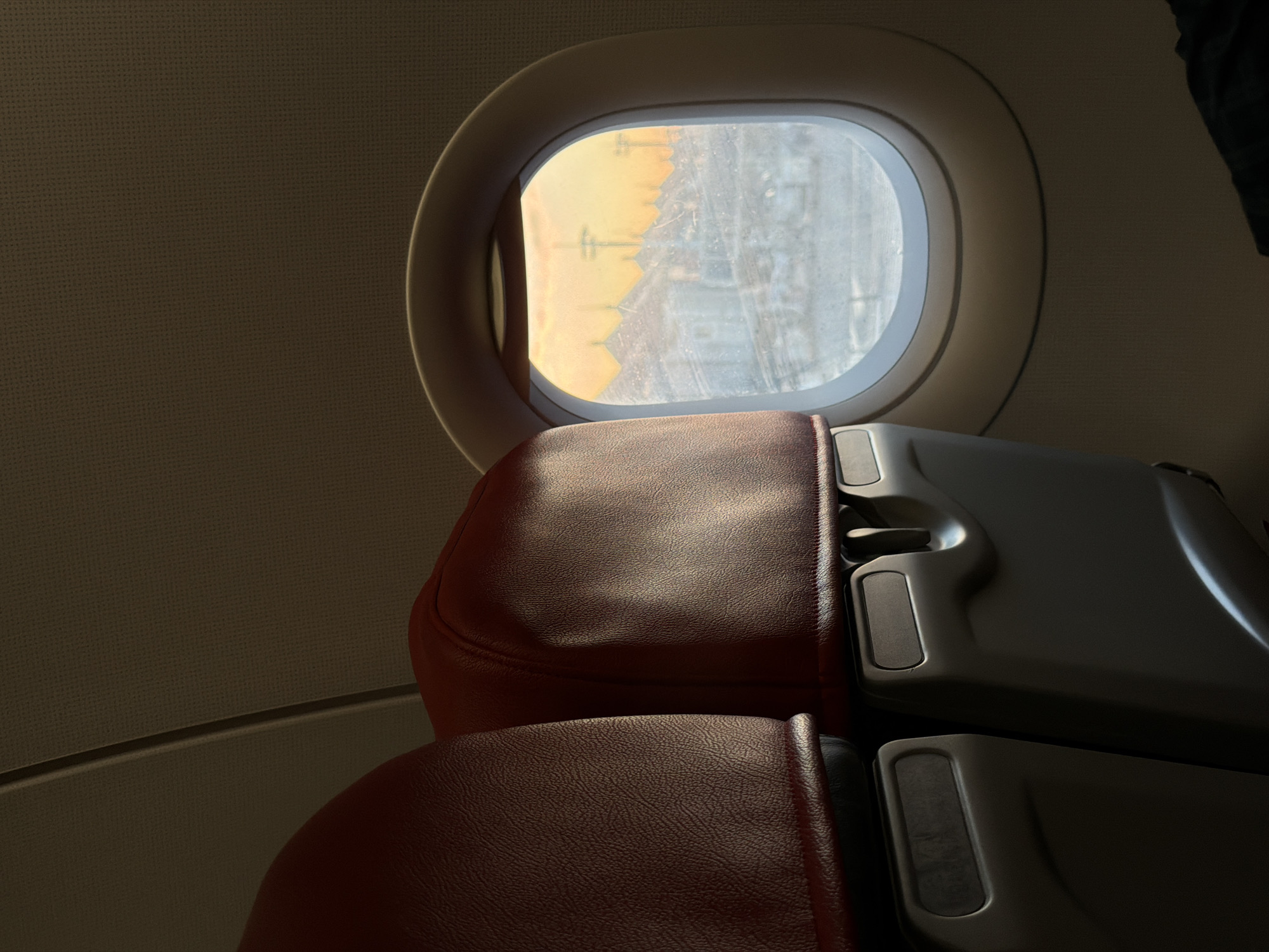 The image shows the interior of an airplane cabin, looking out of a circular window. Two red leather airplane seats are visible in the foreground, positioned to the left of the frame. The window, on the right side, reveals a city skyline at sunset. The sky is a warm orange and yellow, casting a soft light on the buildings below. The architecture appears to be a mix of traditional and modern styles, with several buildings featuring domed roofs and minarets, suggesting a location with Islamic influence. The city below is blanketed in a light layer of frost or snow, indicating a cold climate. The view from the window is slightly obscured by condensation or frost on the glass. There are no people visible within the frame, but the presence of the airplane seats implies travelers. The setting appears to be dusk or dawn, given the color of the sky. The overall scene conveys a sense of travel and the vastness of the world seen from above. Based on the visible architectural elements and the sunset glow, this could be a view of the historical city of Kuta, Indonesia, which is known for its beautiful sunsets and diverse architectural landscape.